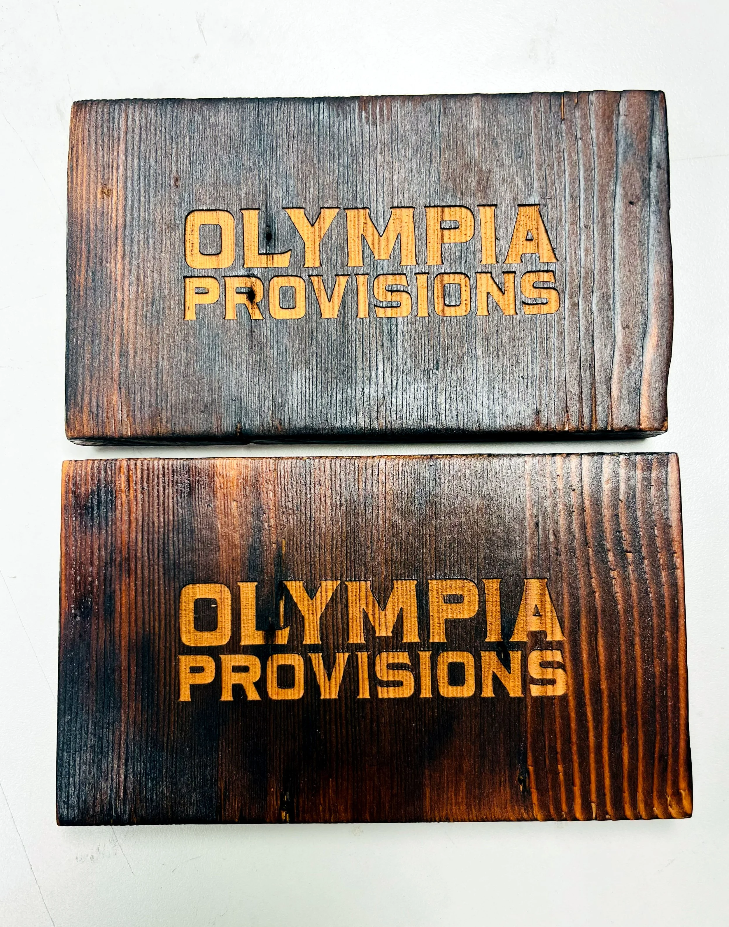 CO2 laser engraved wooden block reading 'OLYMPIC PROVISIONS' with orange highlighted lettering, custom laser engraving by MC Laser Labs.
