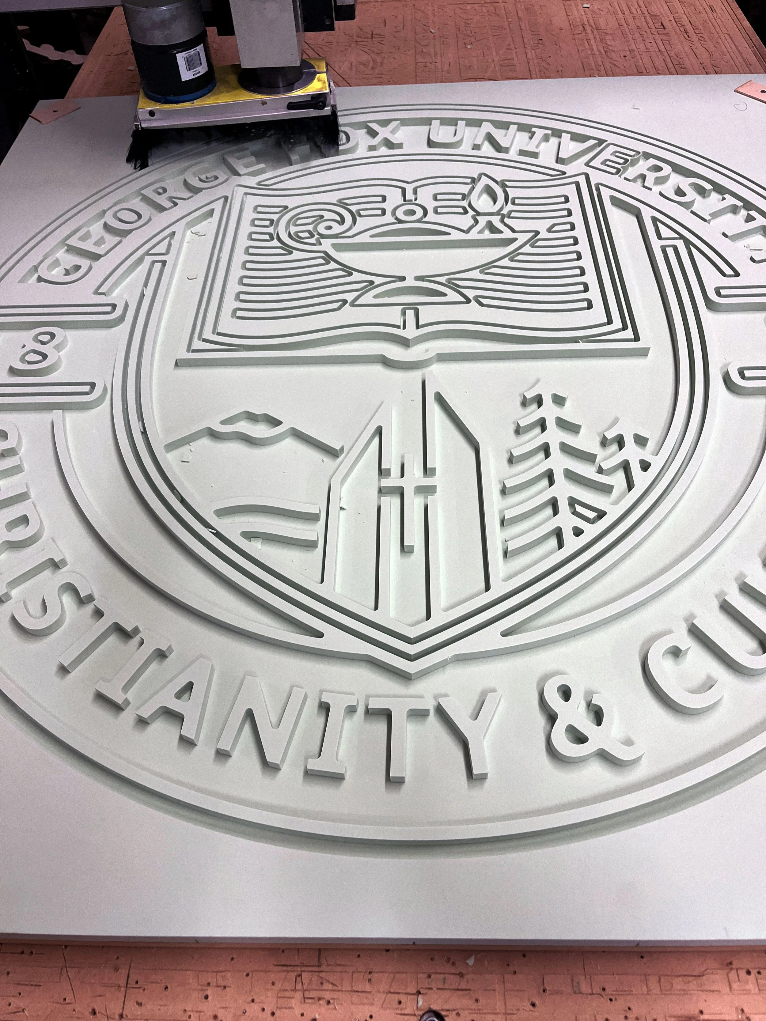A white circular sign with raised letters and symbols, including a book and a degree symbol, with the words 'GEORGE' and 'UNIVERSITY' visible. The sign is on a table with a machine above it, likely for cutting or engraving the sign.