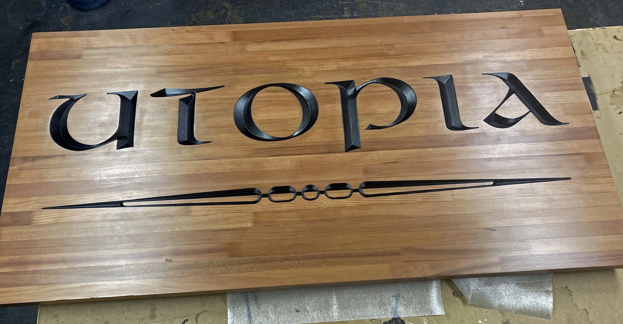 Black 3D letters spelling 'Utopia' on a wooden surface, with a black metal tuning fork or similar tool below the letters.