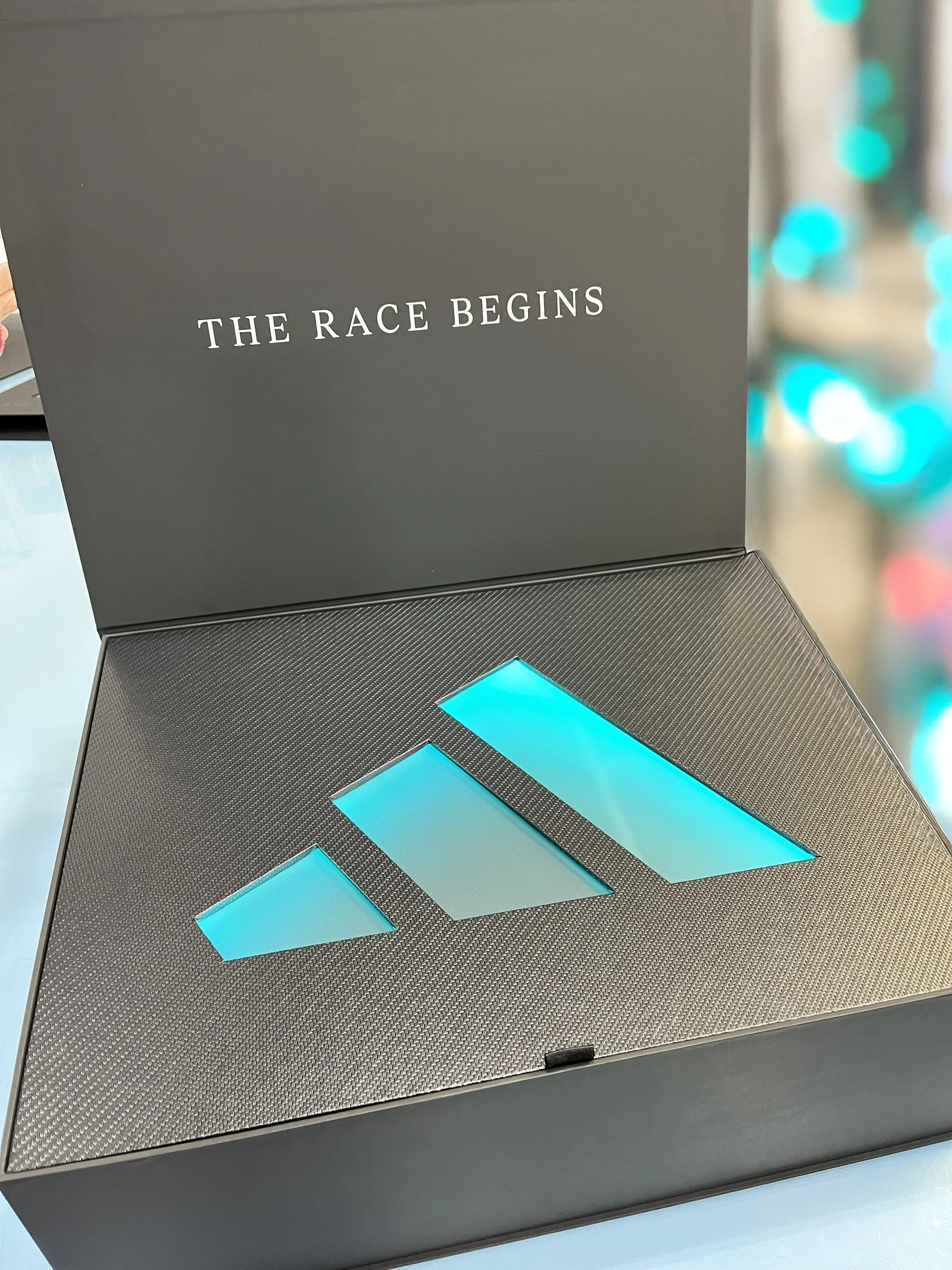 Light-up turned edge seeding kit box for Adidas x Aston Martin F1, reading 'The Race Begins' with illuminated turquoise geometric design, custom fabrication by MC Laser Labs.