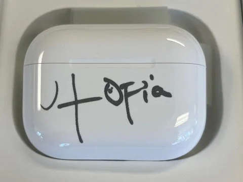 White AirPods case with black handwritten signature that says 'Utopia'