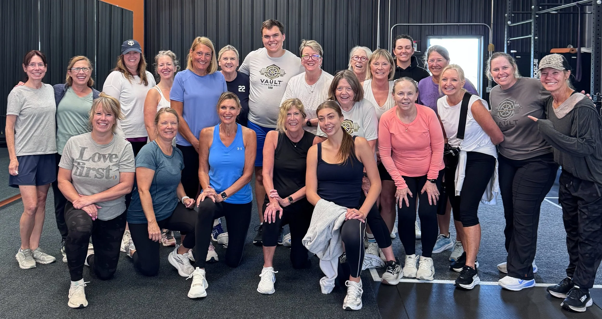 Group of people in workout clothes inside a gym, smiling and posing for a group photo, with exercise equipment visible in the background.