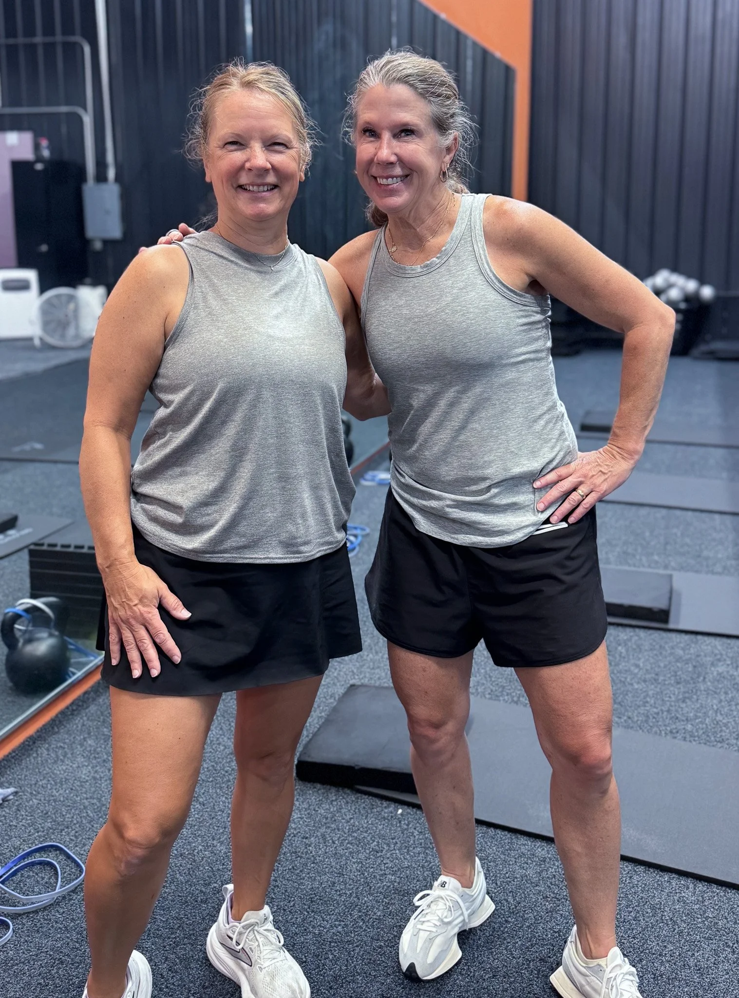 Two smiling women in workout clothes standing in a gym, with fitness equipment in the background.