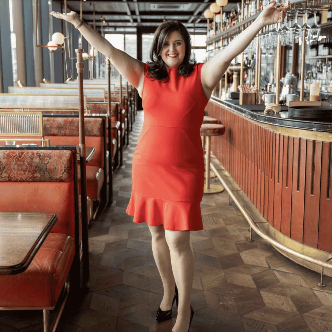 A woman in a red dress and black heels standing with arms raised inside a retro-style restaurant or bar with red booth seating and a wooden bar counter.