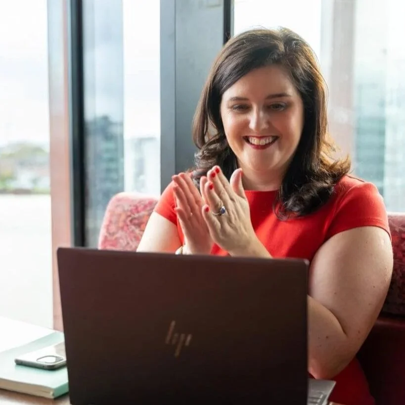 A woman sitting at a desk, smiling and clapping while looking at her laptop. She has dark hair, is wearing a red dress, and has a smartphone and a notebook on the desk.