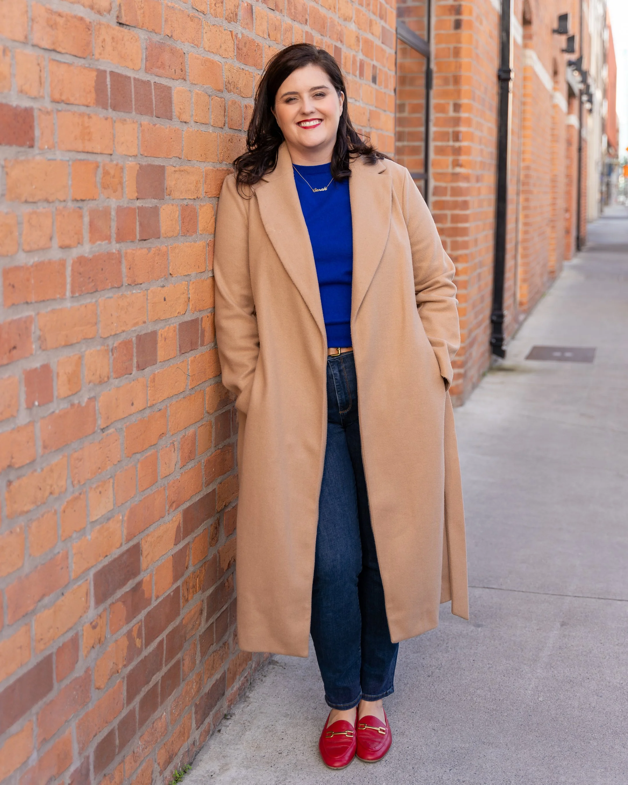 A woman with dark brown hair, wearing a beige coat, blue sweater, and red loafers, stands against a brick wall on a city sidewalk, smiling.