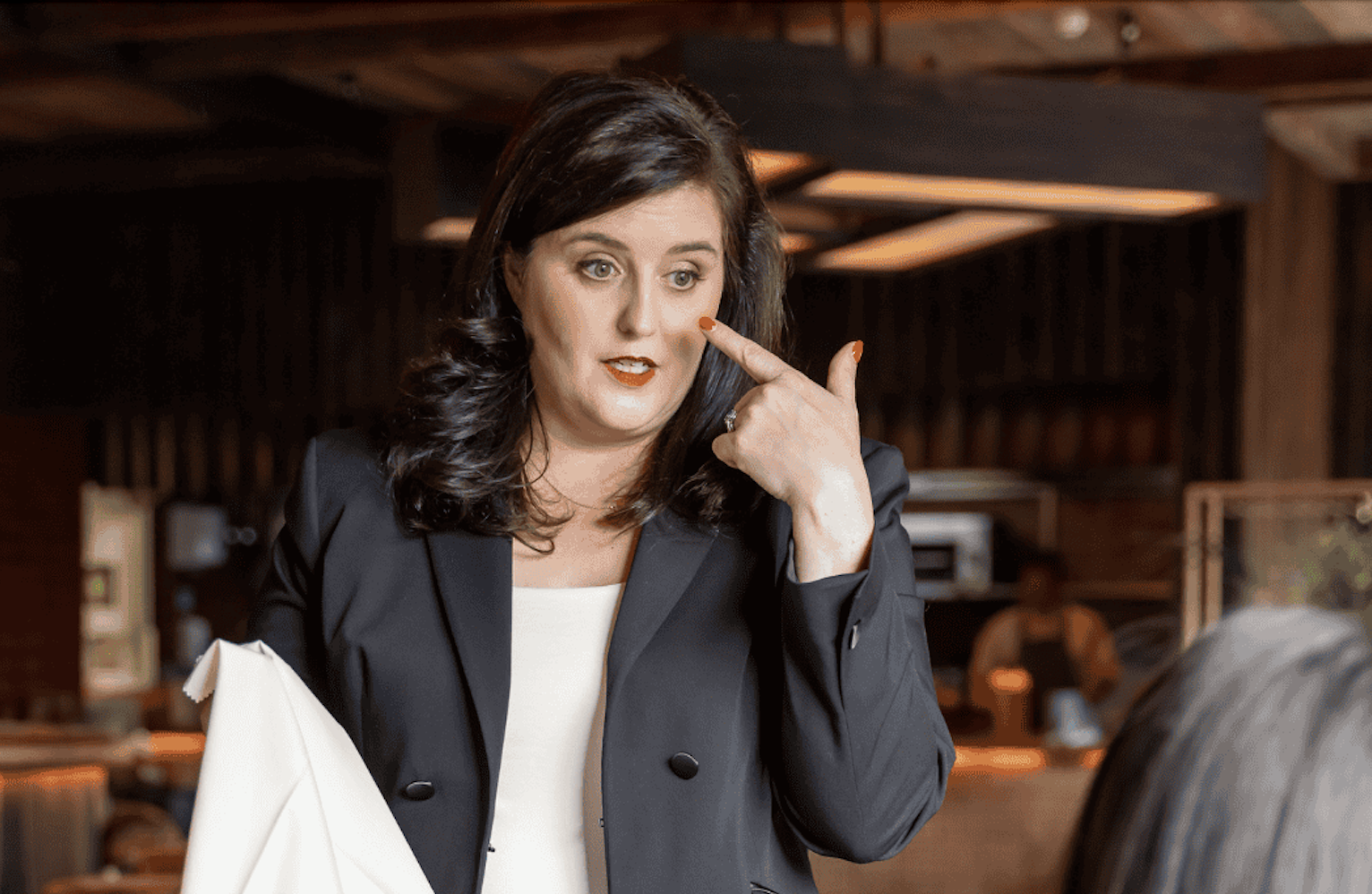 Woman with dark hair wearing a black blazer and white top, gesturing her face with her finger, indoors with warm wooden decor.