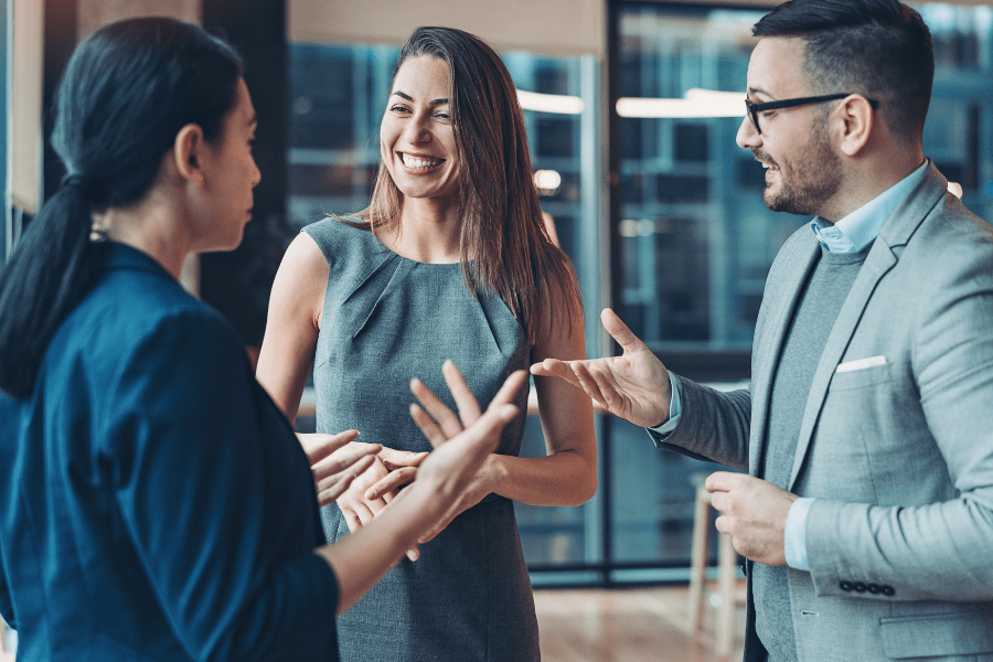 Three professionals having a friendly conversation in an office setting, smiling and gesturing with their hands.
