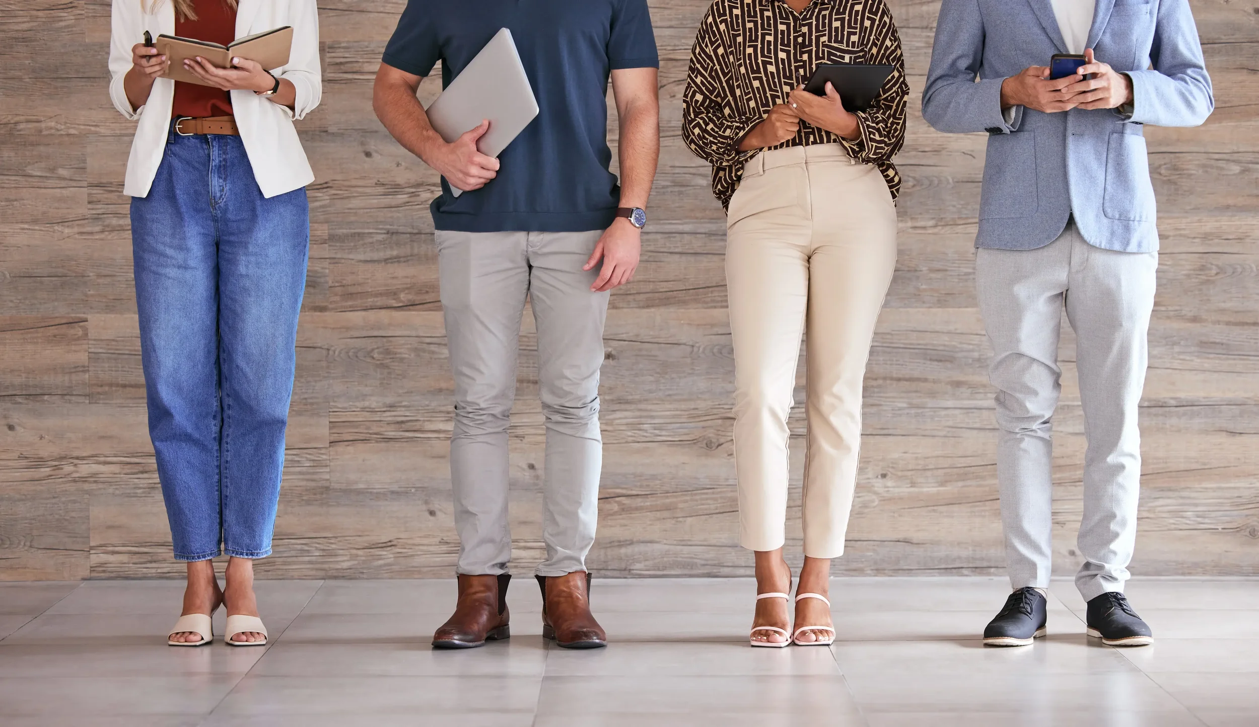 Four people standing side by side, focusing on their legs and feet, against a wooden wall background. They are dressed in casual and business casual attire, holding various electronic devices.
