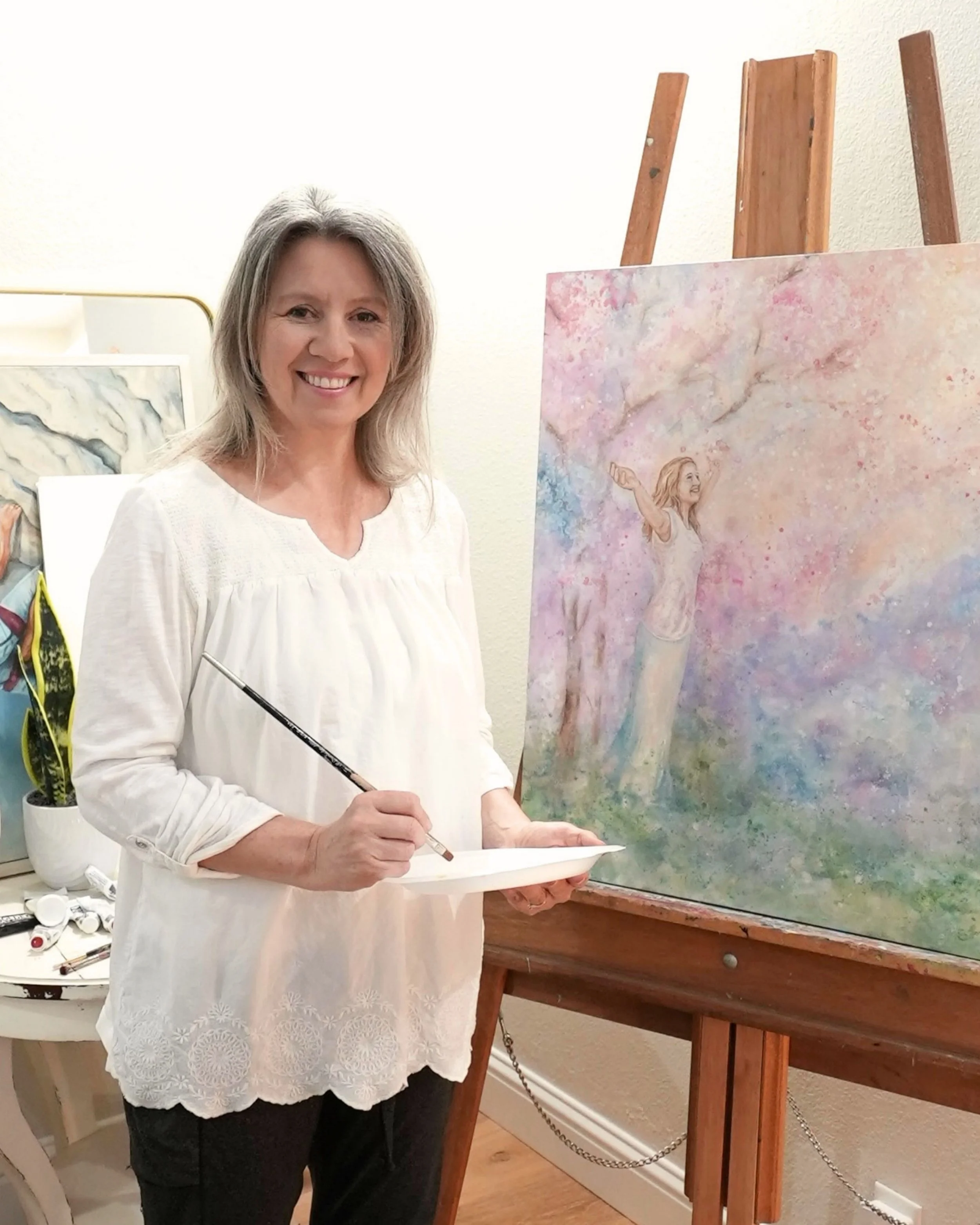 Sheira Dawn standing beside one of her original paintings in her studio, holding a paintbrush and palette.