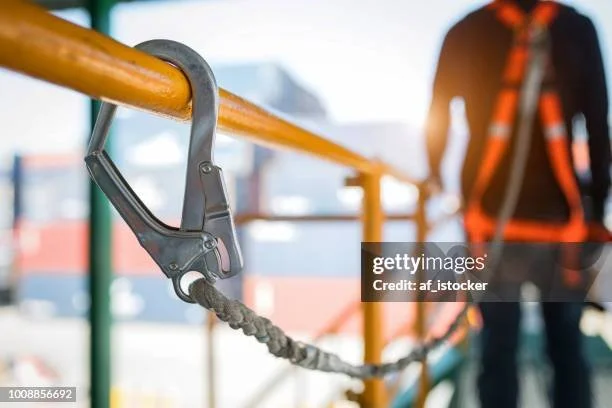 Une personne avec un casque de sécurité, une veste orange et un harnais de sécurité, à côté d'une barrière jaune en chantier.