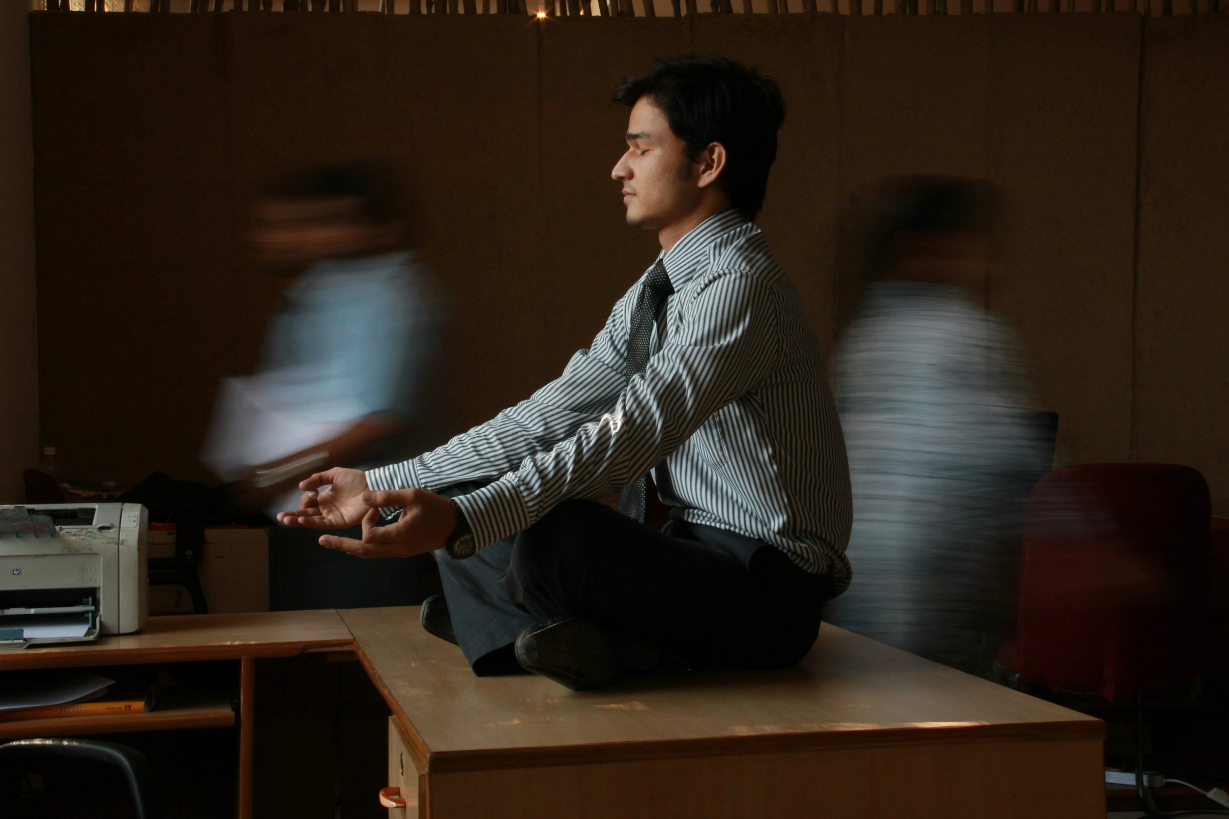 Un homme en costume sombre, assis en position de méditation, effectuant une posture de yoga ou de pleine conscience dans un bureau, avec deux collègues flous qui passent derrière lui.