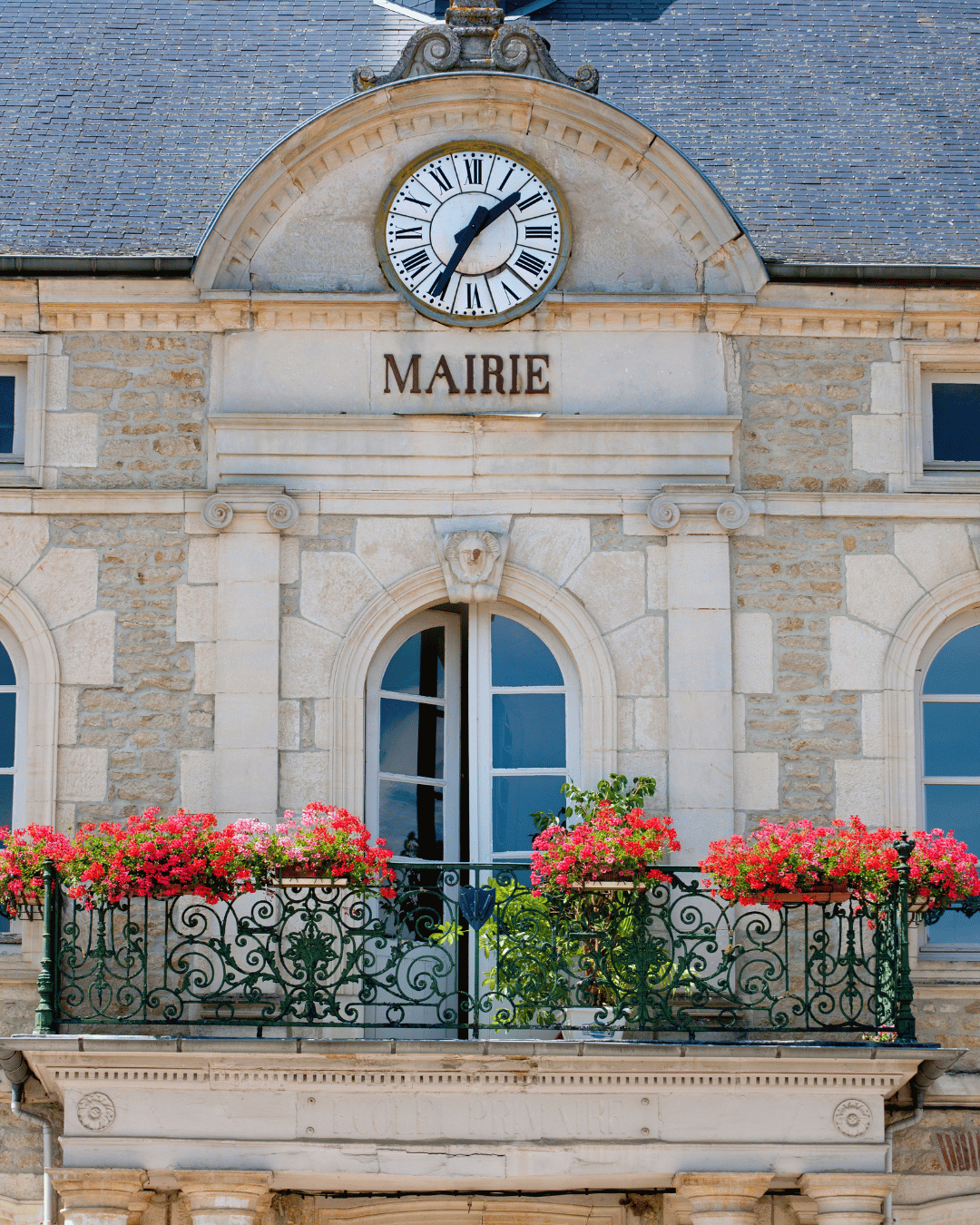 Bâtiment en pierre avec horloge, inscription 'MAIRIE', balcon avec fleurs roses et plantes vertes.