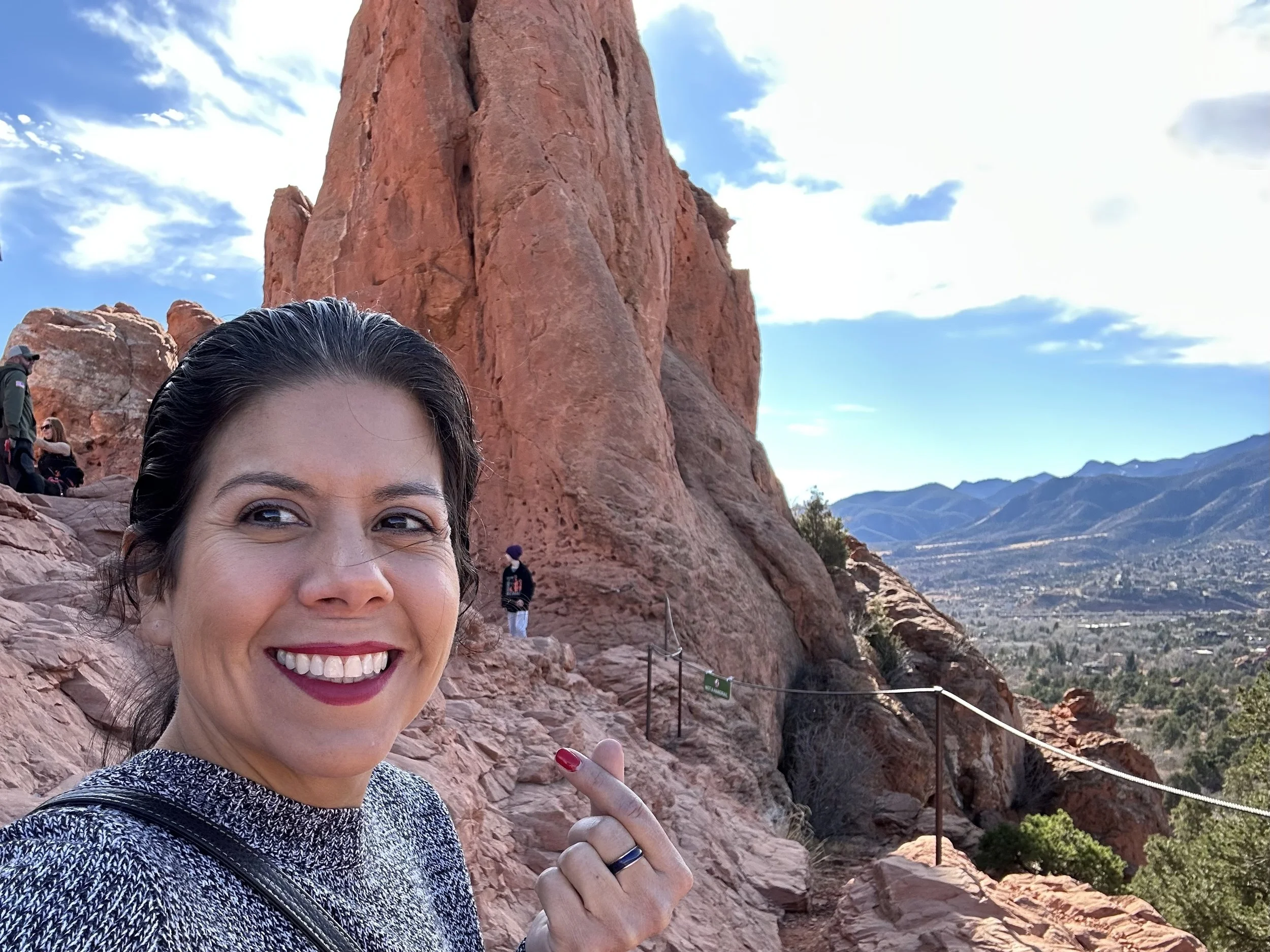 A woman smiling at the camera with a large red rock formation and a scenic mountain valley in the background.