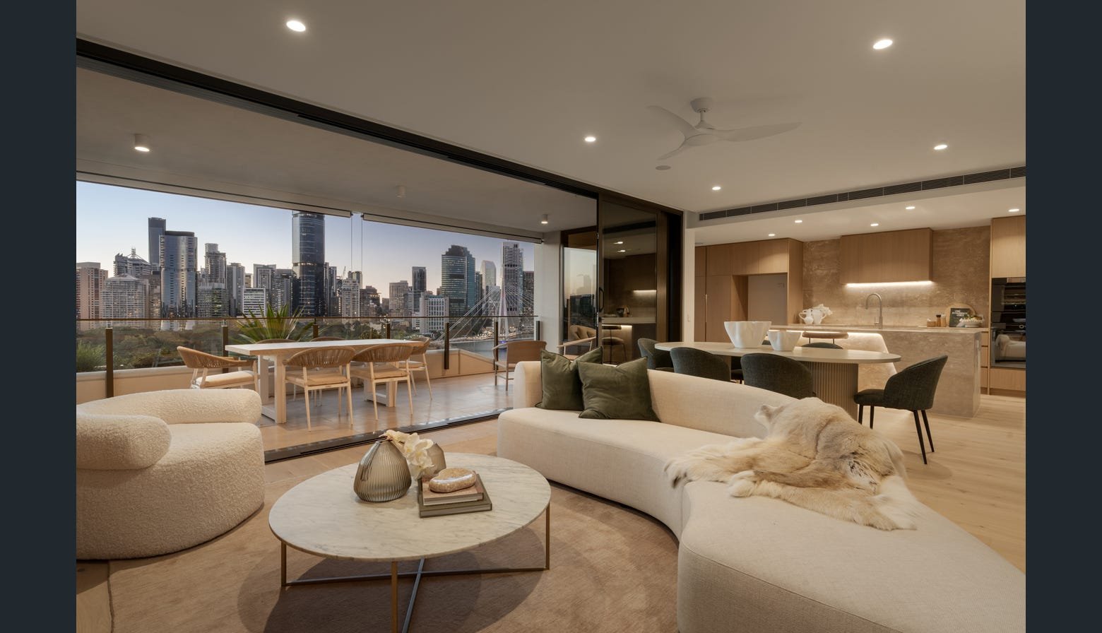 Modern city skyline view from a spacious apartment with a living room, dining area, and balcony. The room features neutral tones, a large white curved sofa with pillows, a round marble coffee table with decorative items, and a dog lying on the sofa.