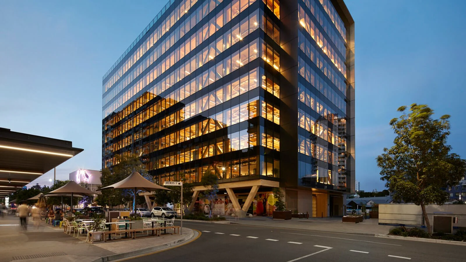 A modern glass office building illuminated at dusk with visible interior lighting, surrounded by a street, trees, and outdoor seating.