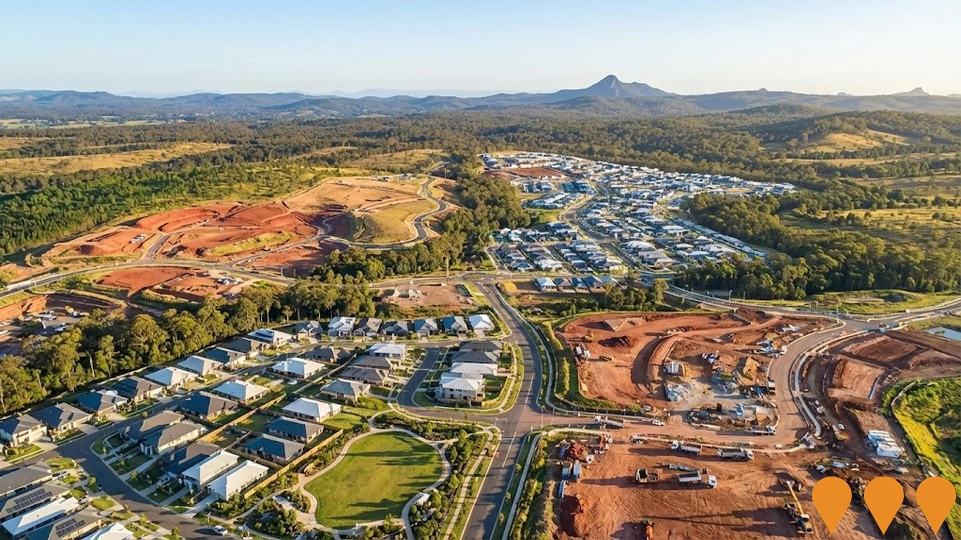 Aerial view of a neighborhood with white houses, roads, and green lawns, next to a large construction site with cleared land, dirt roads, and construction vehicles on hilly terrain, with a forested landscape and mountains in the background.
