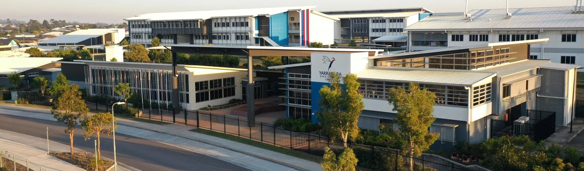 A modern school building with the sign 'Yarra Ranges State Secondary College' surrounded by trees and a schoolyard, with a nearby street in the foreground.