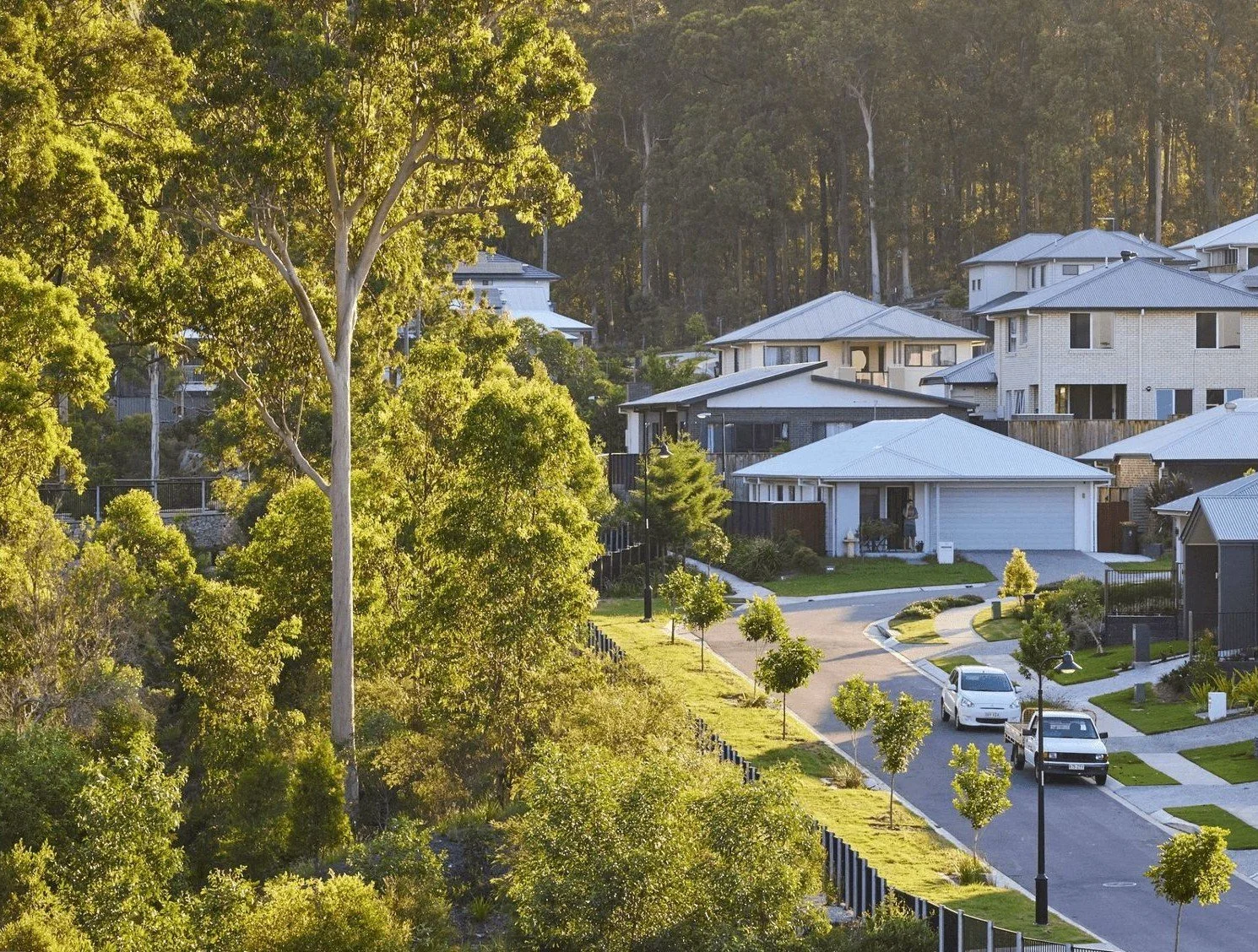 Residential neighborhood with modern houses, trees, and a curved driveway with parked cars, in a hilly area.