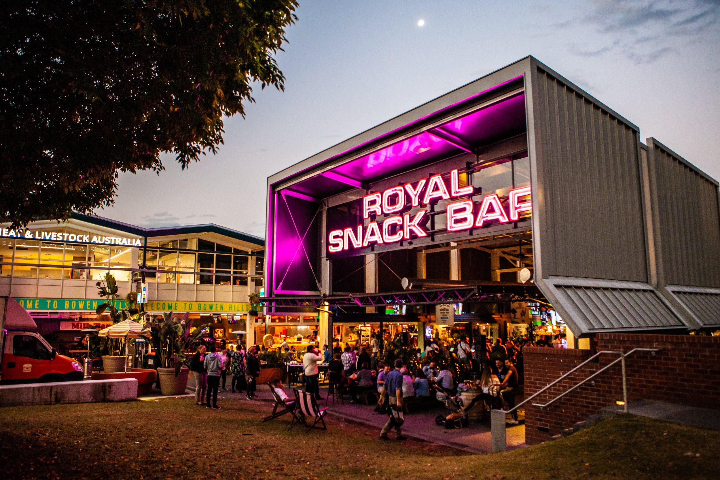 Night scene of a crowded outdoor area with a modern food or snack stand that has a vibrant pink neon sign reading 'Royal Snack Bar'. There are people sitting and standing, some under umbrellas, with trees and a building in the background. The sky is dark with the moon visible.