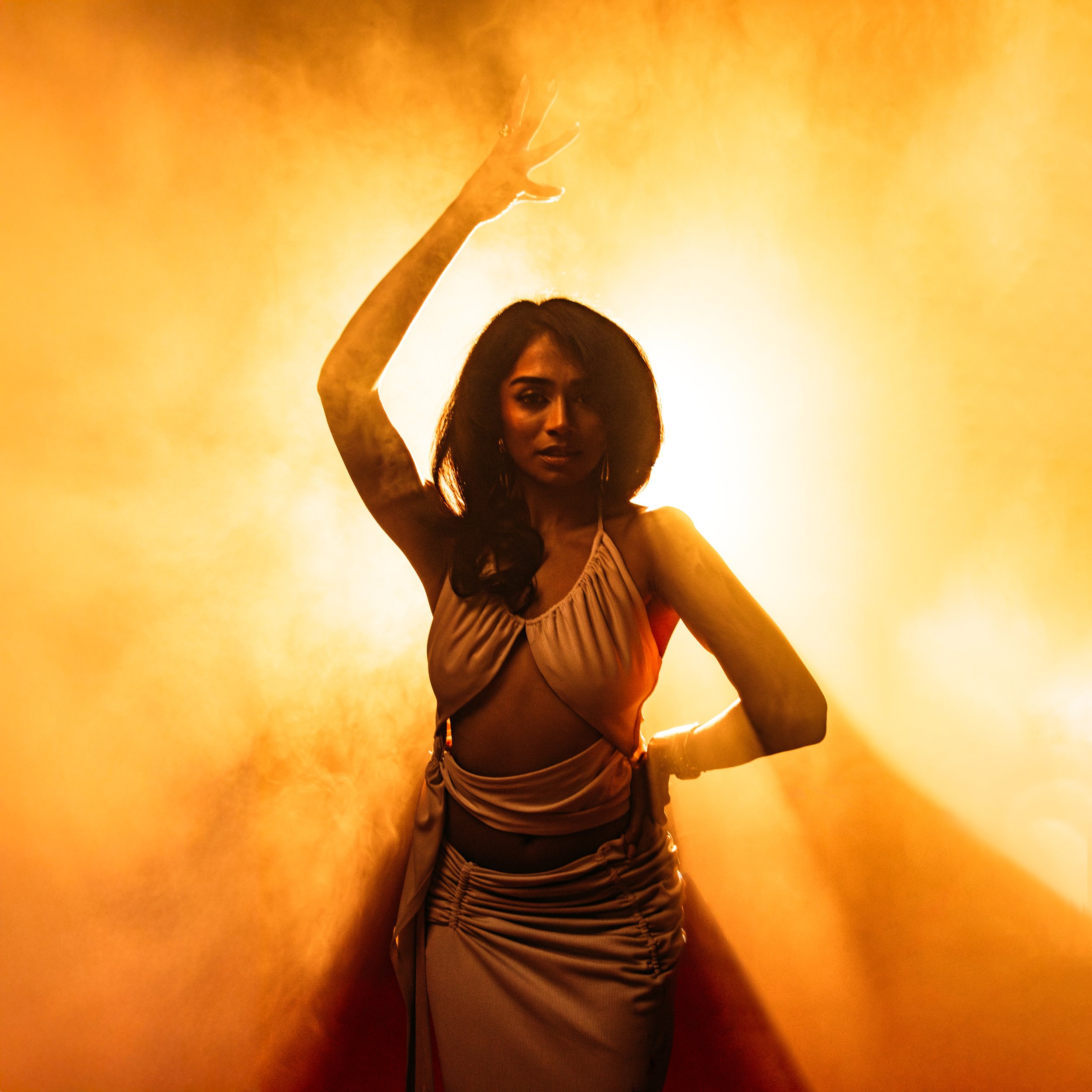 A woman with dark hair and a draped dress striking a pose against a bright yellow-orange background.