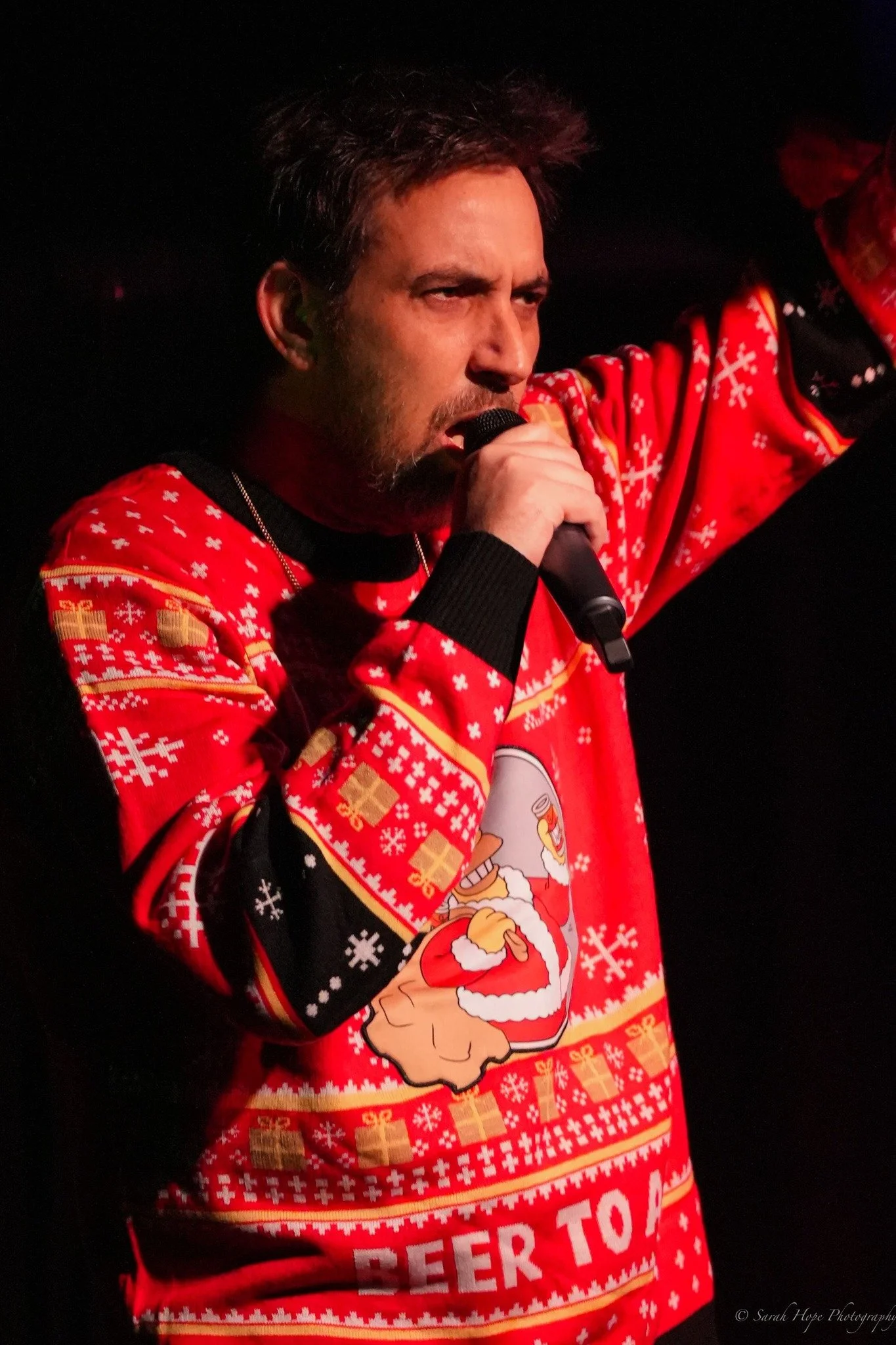 Photo of Wild Oscah hosting and performing at The Mixmas at The Carleton Dec. 5th 2025 wearing a bright red Christmas sweater, deep contrast photo taken by Sarah Hope Photography