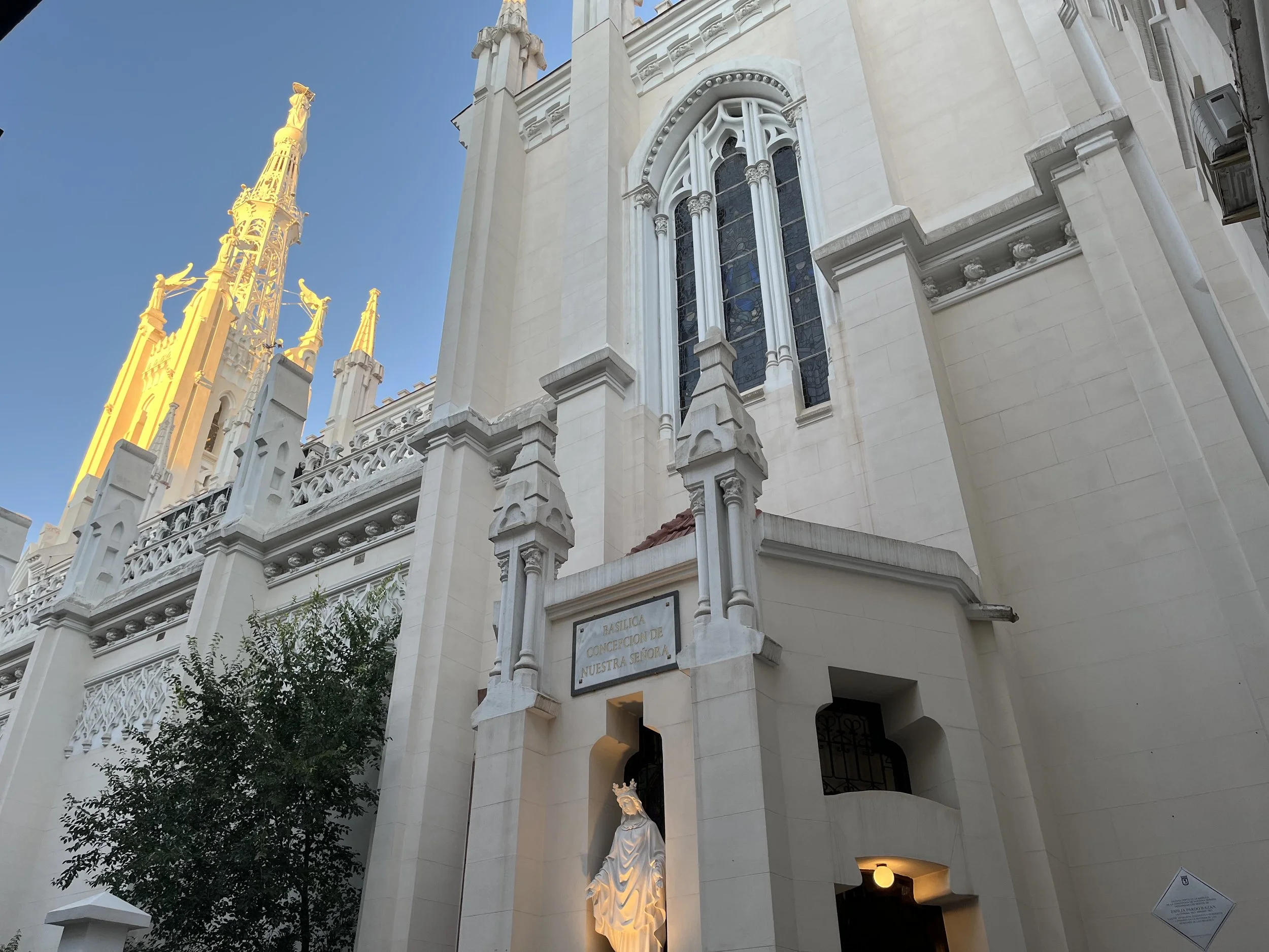 The exterior of a Gothic-style church building in Madrid, Spain, with detailed white stone architecture, with a large stained glass window and tall spires, under a clear blue sky.