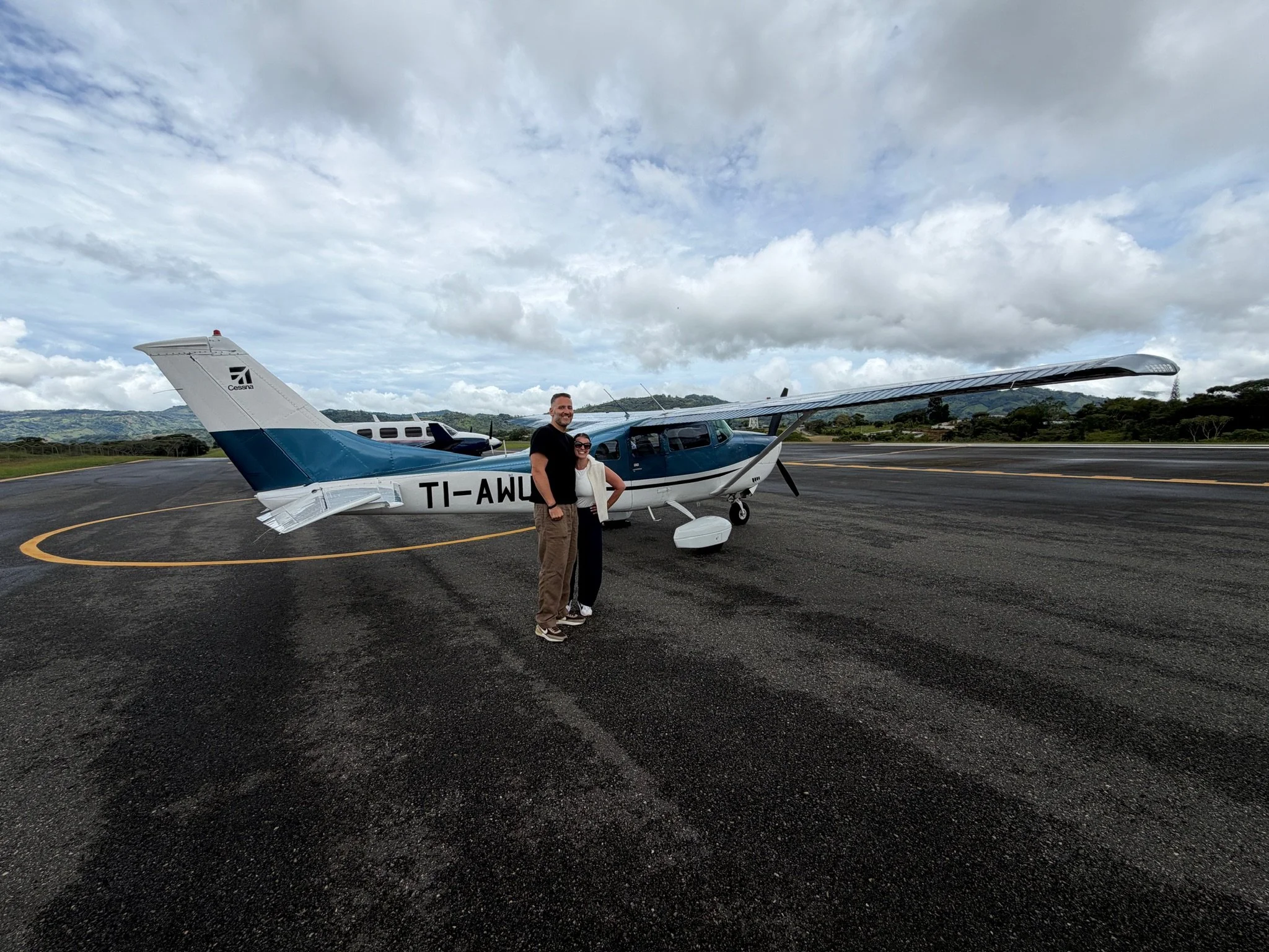 Two smiling people, a man and a woman, standing in front of a small blue and white airplane on an airport tarmac with cloudy skies and distant hills in the background.