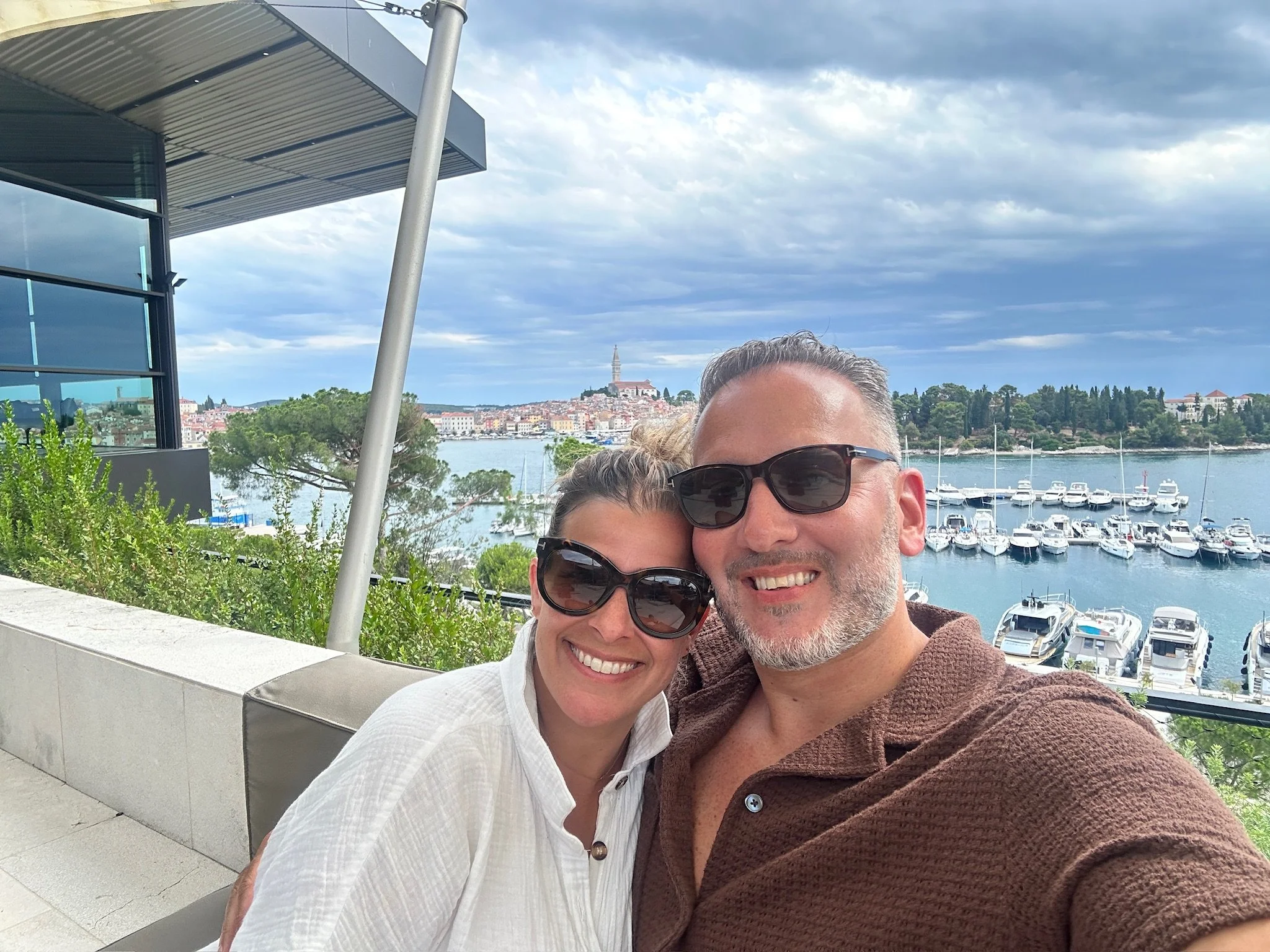 A smiling couple taking a selfie outdoors with a marina, boats, and a hillside town in the background.