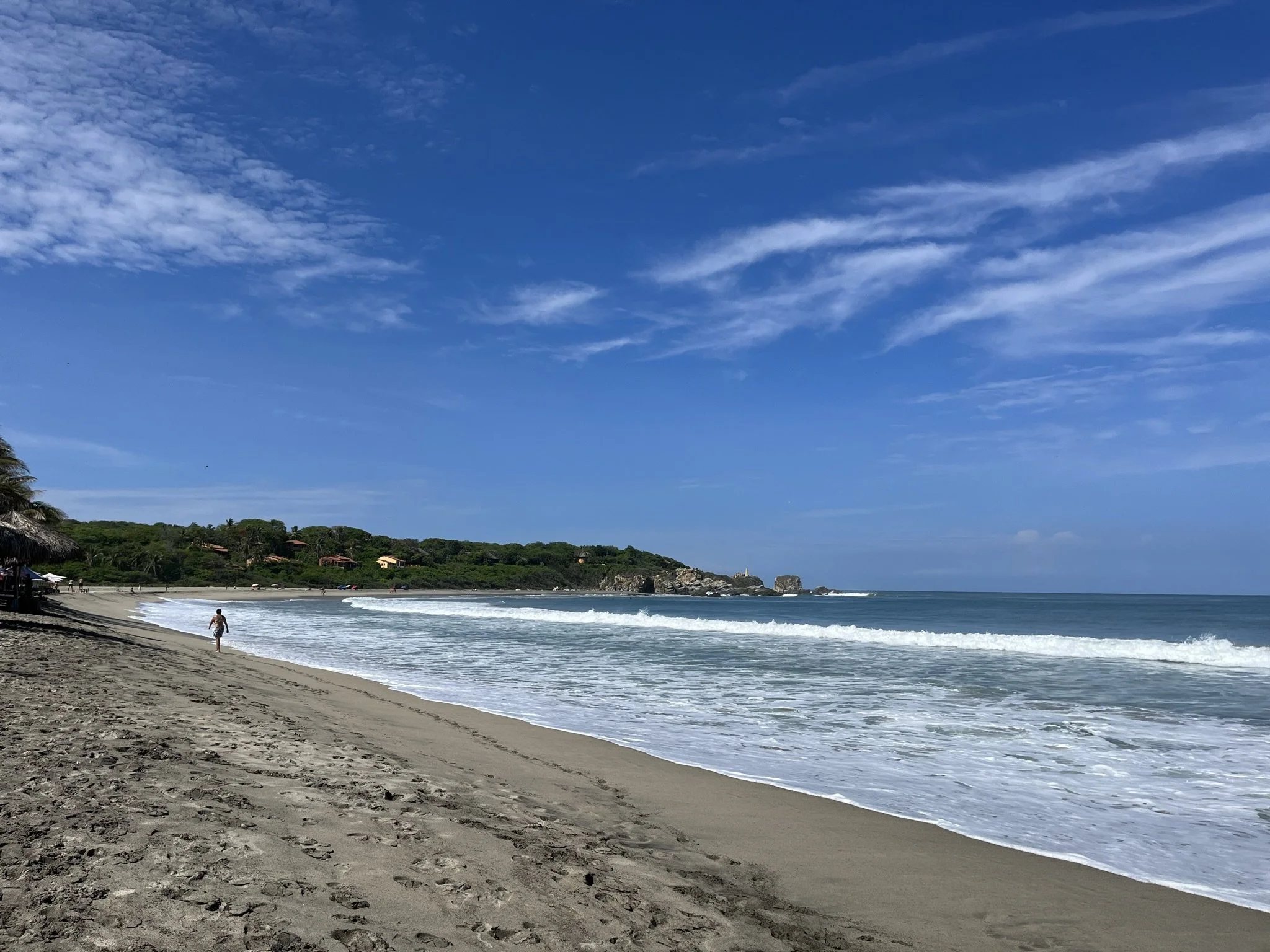 The sandy beach with ocean waves, tropical greenery, and houses on a hillside under a blue sky with scattered clouds in La Punta, Puerto Escondido, Mexico.