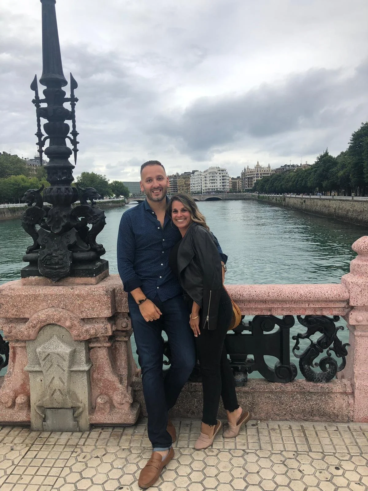 A couple smiling and embracing on a bridge over a river, with city buildings and cloudy sky in the background.