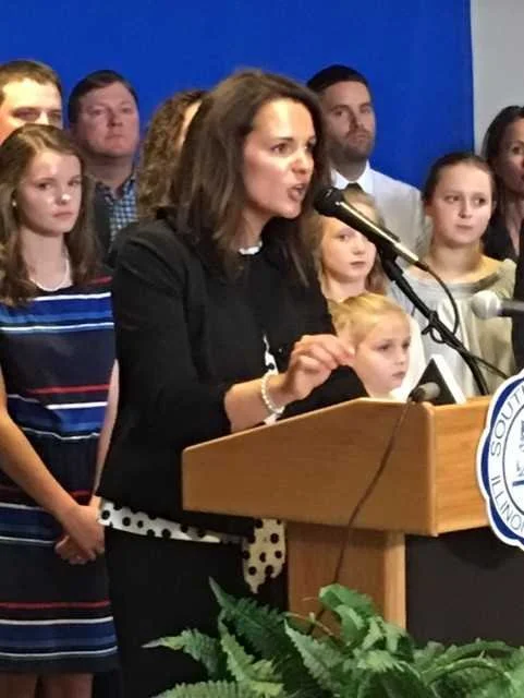 A woman speaking at a podium during a press conference or public event, with a group of children and adults standing behind her.