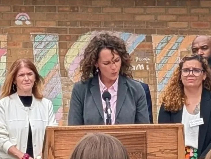 A woman speaking at a podium during a press conference or public event, with three other women standing behind her against a brick wall with colorful chalk drawings.