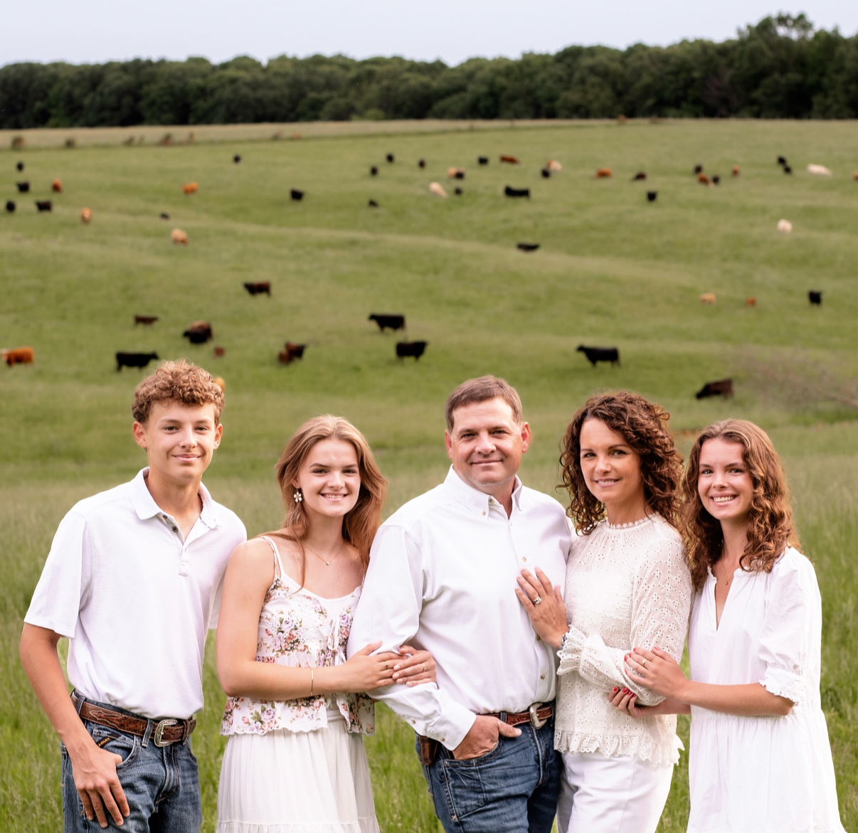 Family of five standing together in a grassy field with cows grazing on a hill in the background.