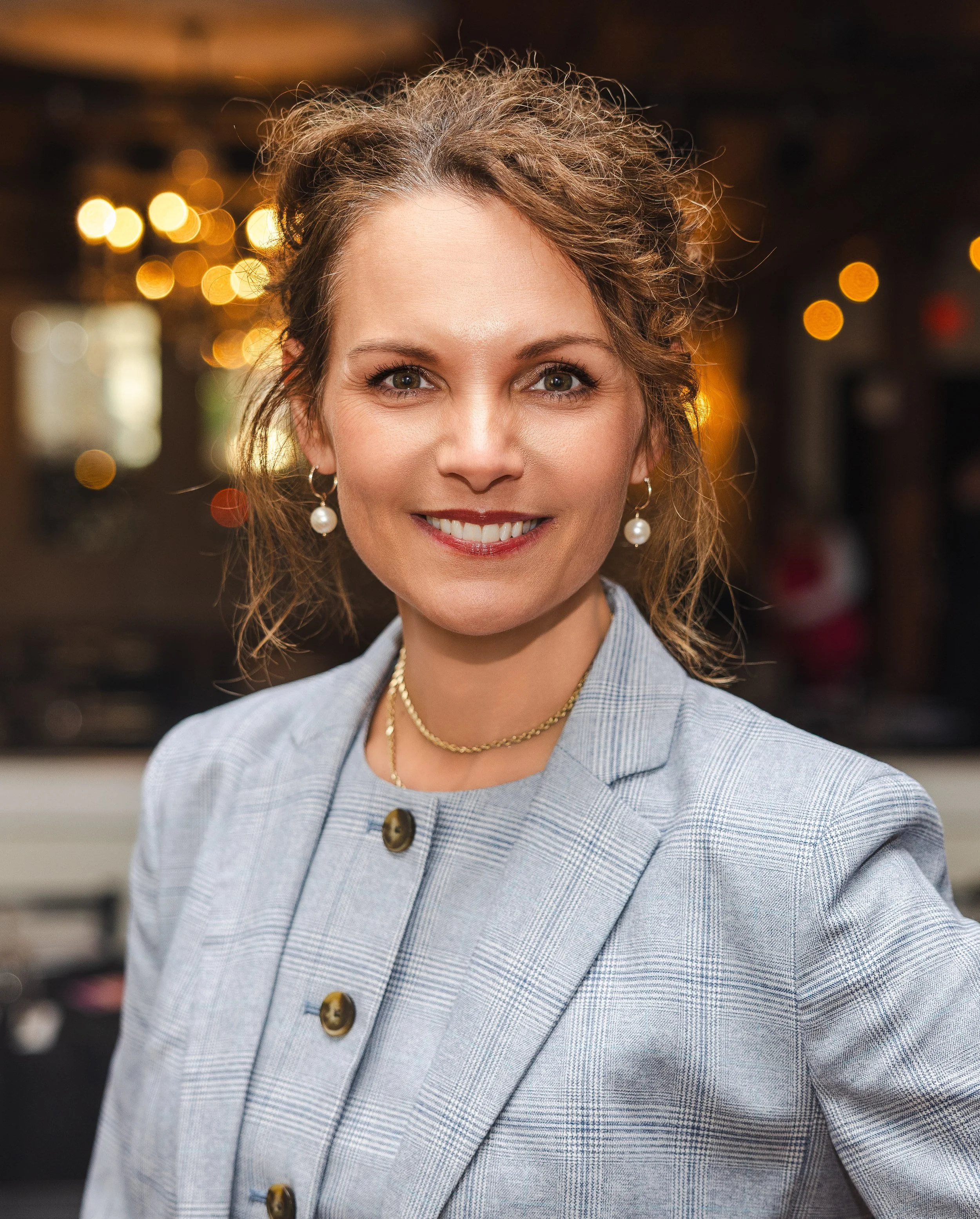 A woman with curly light brown hair smiling confidently in an indoor setting with warm lighting and shelves in the background.