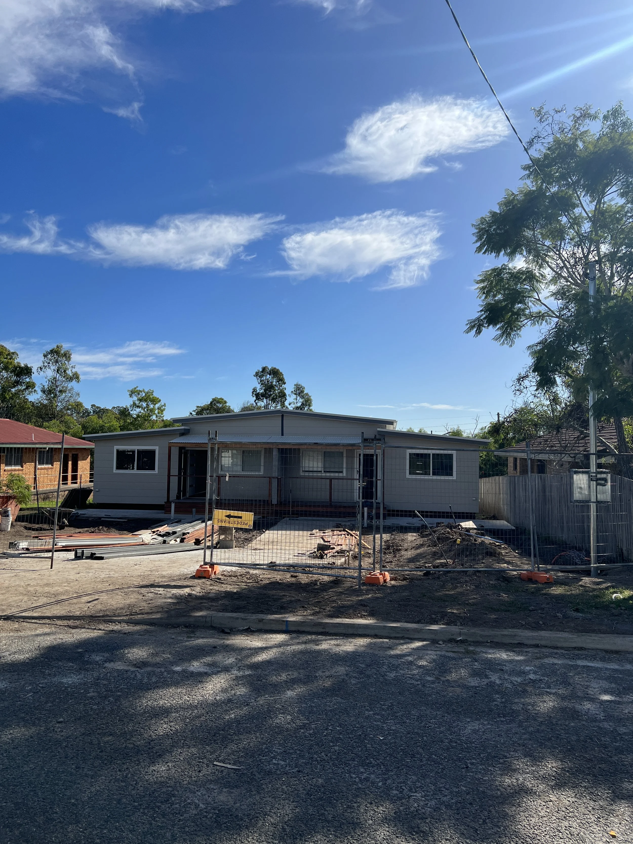 A single-story house under construction, fenced off, with tools and materials in front under a blue sky with scattered clouds.