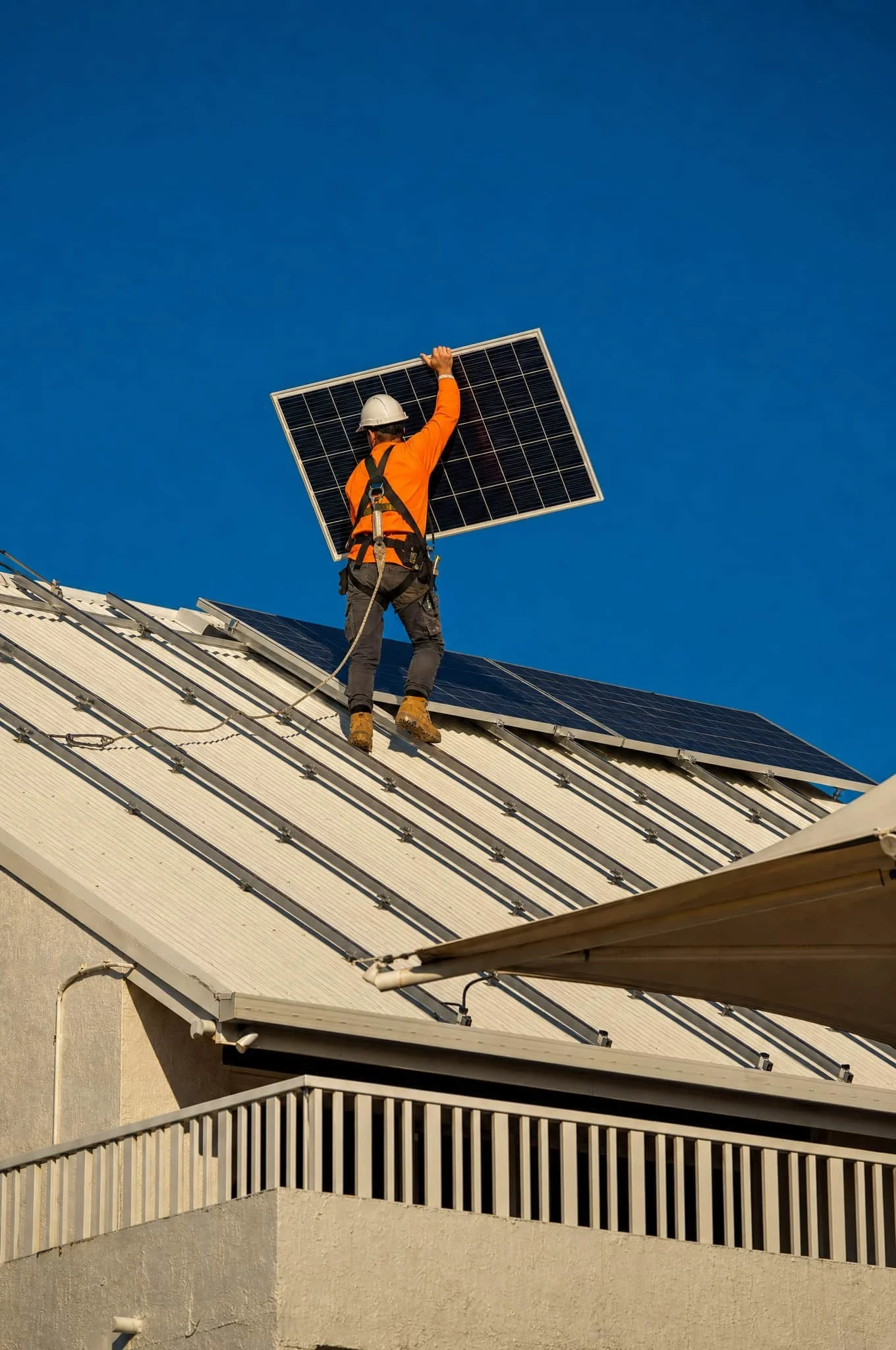 A worker in orange safety gear and a white helmet installing solar panels on a house roof against a clear blue sky.