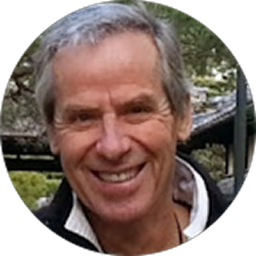 A smiling older man with gray hair outdoors, with trees and possibly a building in the background.