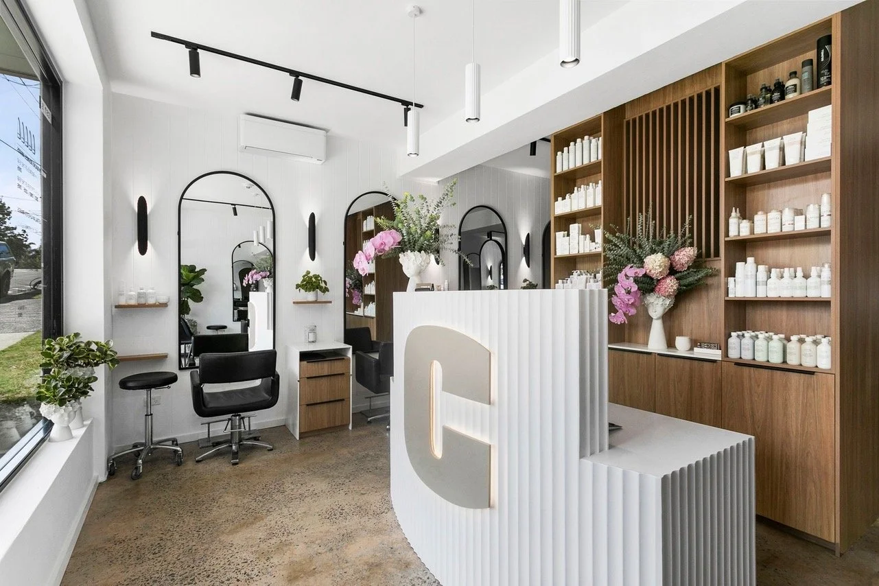Modern salon interior with mirrors, shelves of products, fresh flowers, styling chairs, and a white reception desk with a large C.