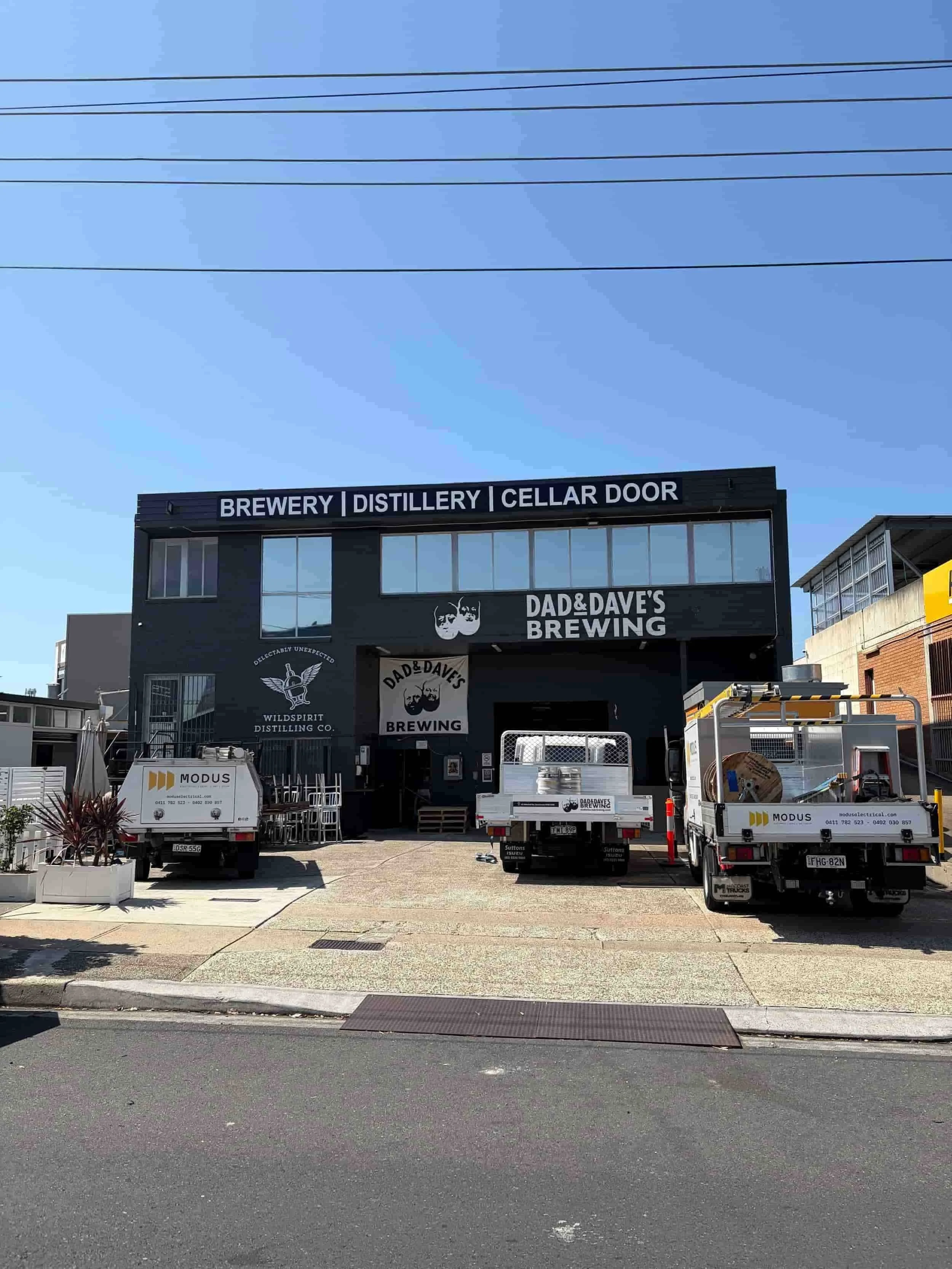 Industrial building with Dad & Daves Brewing signage, two trucks parked in front, and clear blue sky above.