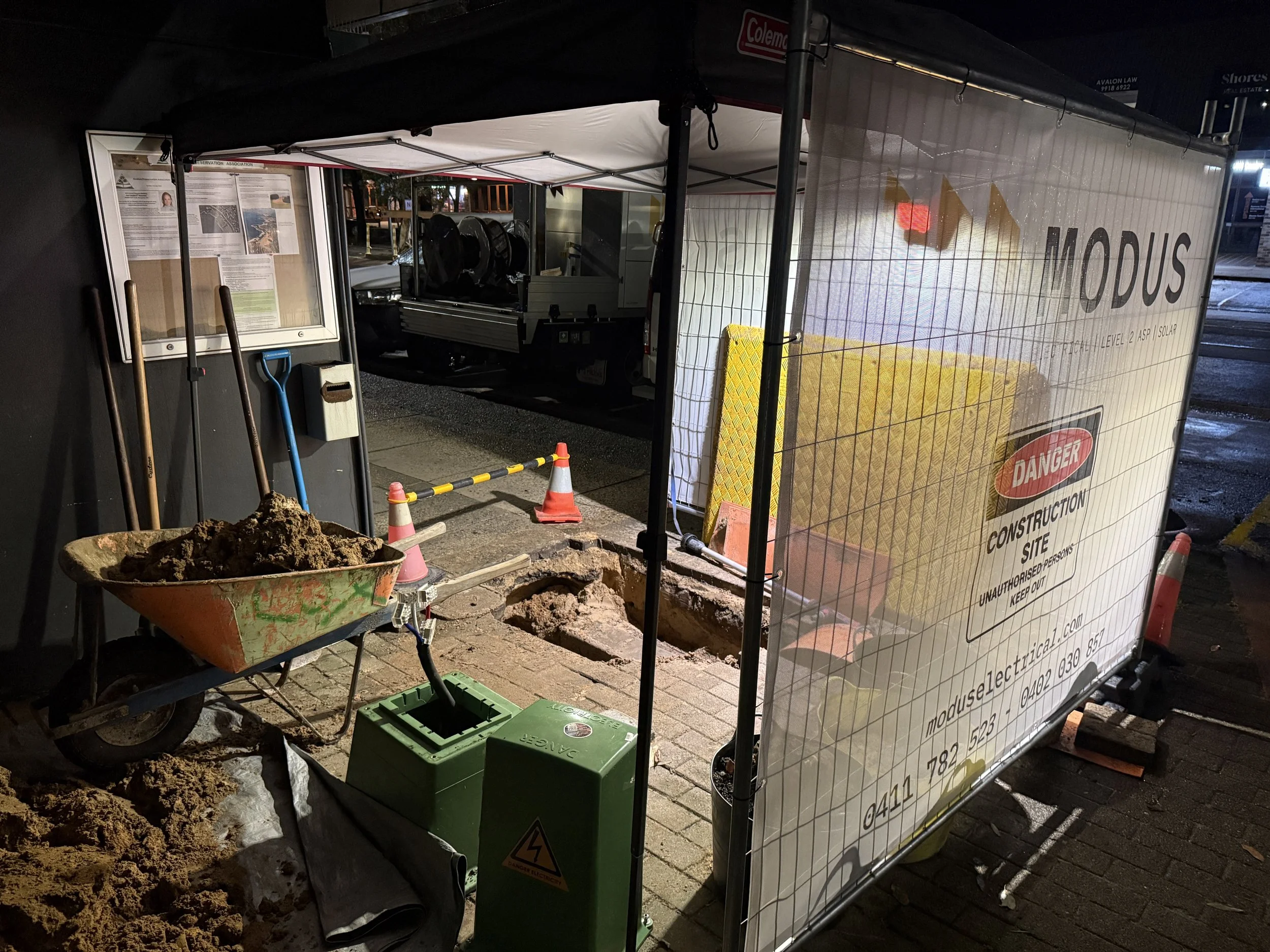 Night scene of a construction site with a fenced-off area, a wheelbarrow filled with dirt, a green generator, and construction cones on a brick sidewalk.