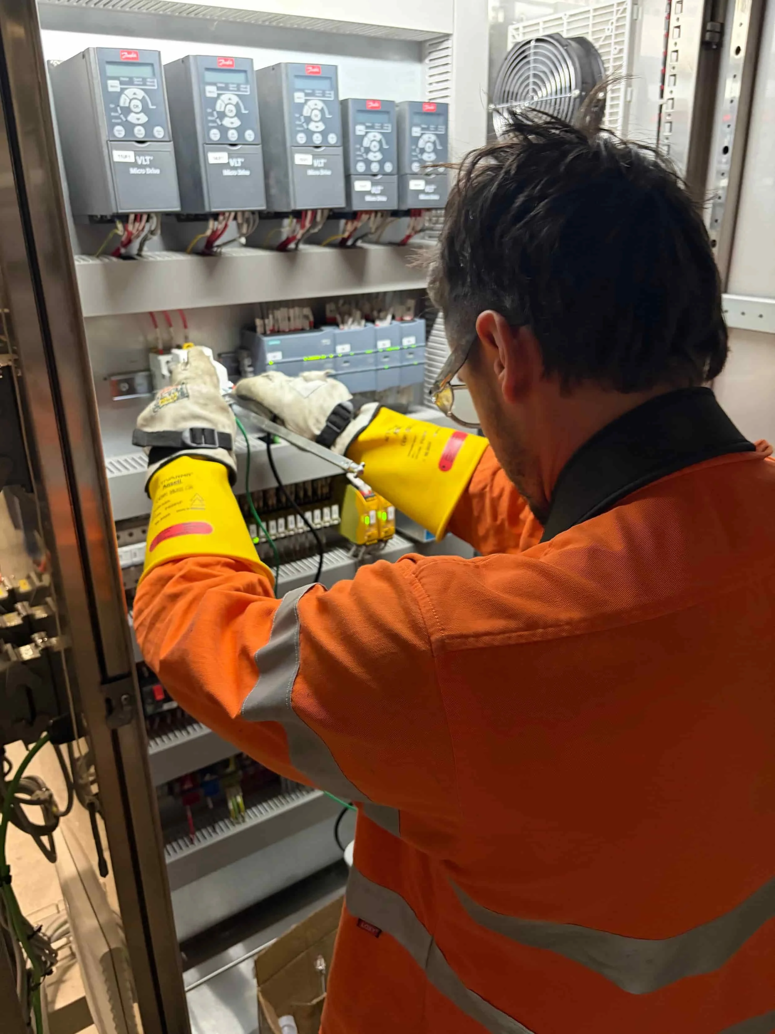 A technician wearing orange protective gloves and safety glasses working on an electrical panel with multiple circuit breakers in a utility room.