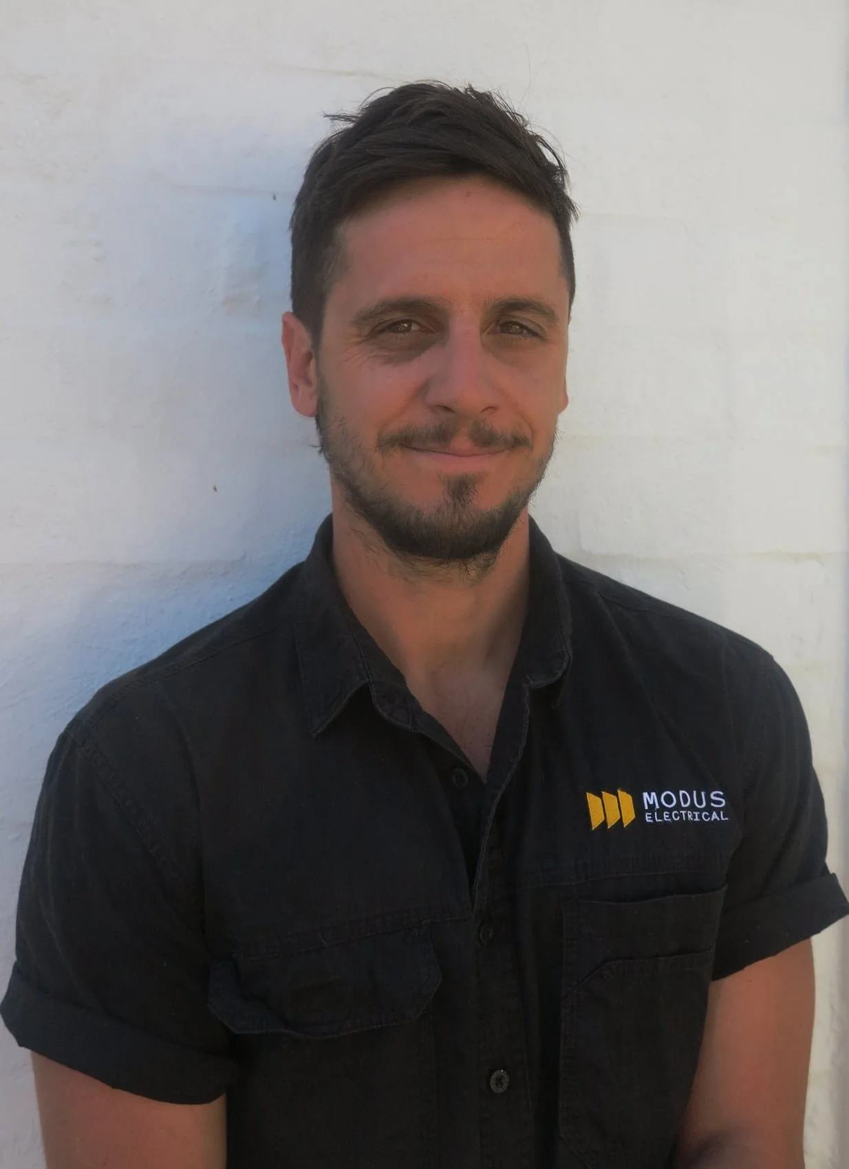 A man with dark hair and facial hair smiling, wearing a black shirt with a logo that says Modus Electrical, standing against a white wall.