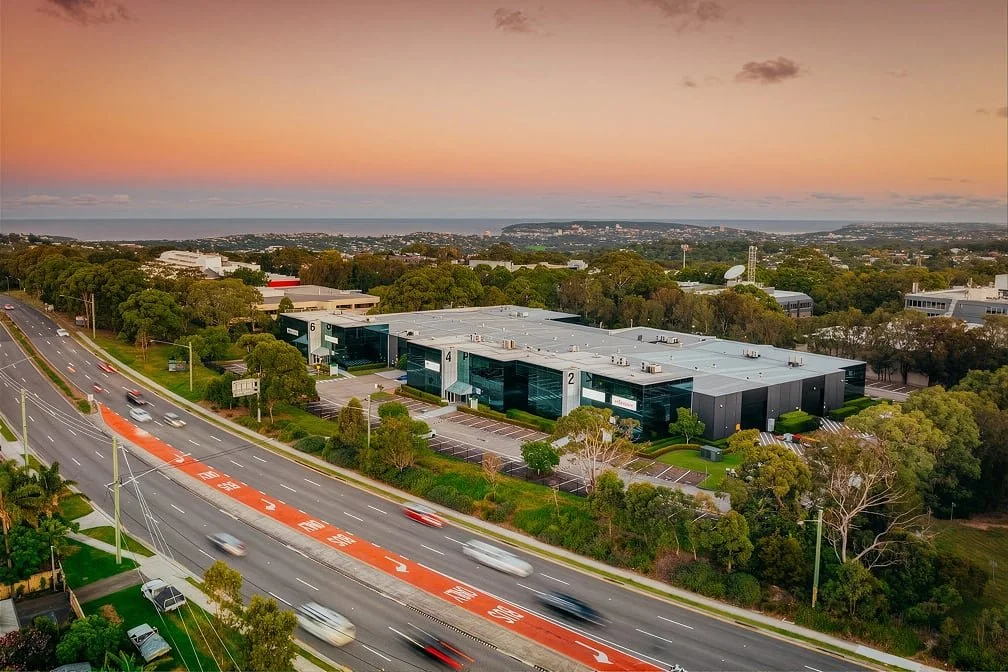 Aerial view of a modern office complex by a busy road with cars, surrounded by trees at sunset.