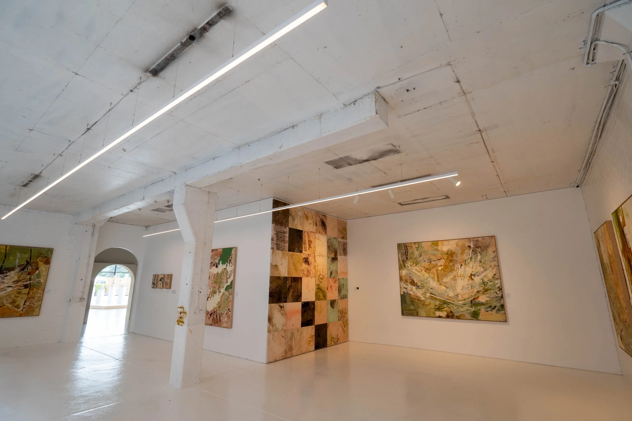 Interior view of an art gallery featuring a large wall with numerous paintings arranged in a grid pattern. The ceiling has exposed pipes and a white beam, with modern linear lighting. There is an arched doorway with natural light coming through, illuminating the white floor.