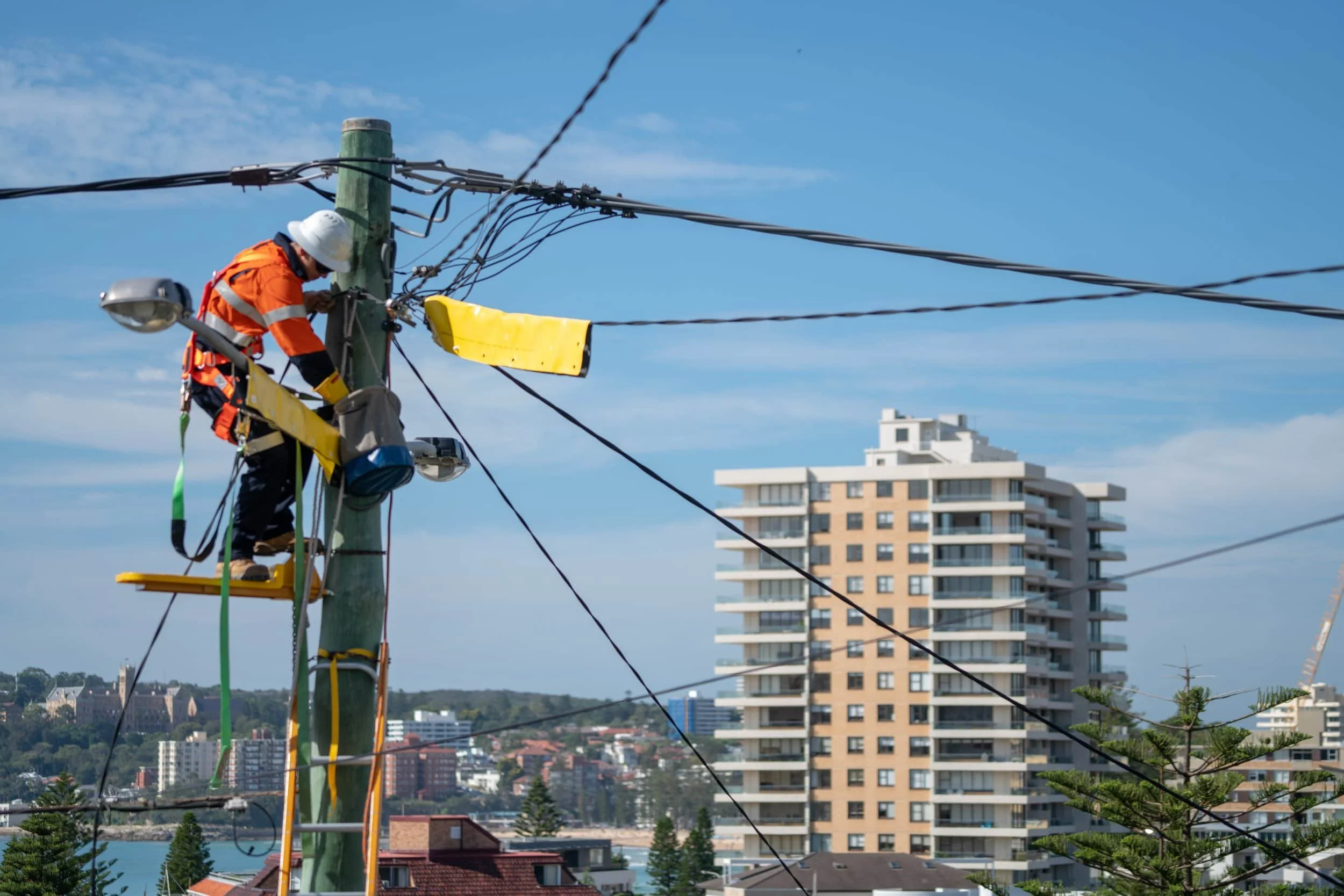 A worker in safety gear repairing or maintaining overhead electrical wires on a utility pole in an urban area with high-rise buildings and residential houses, under a blue sky.