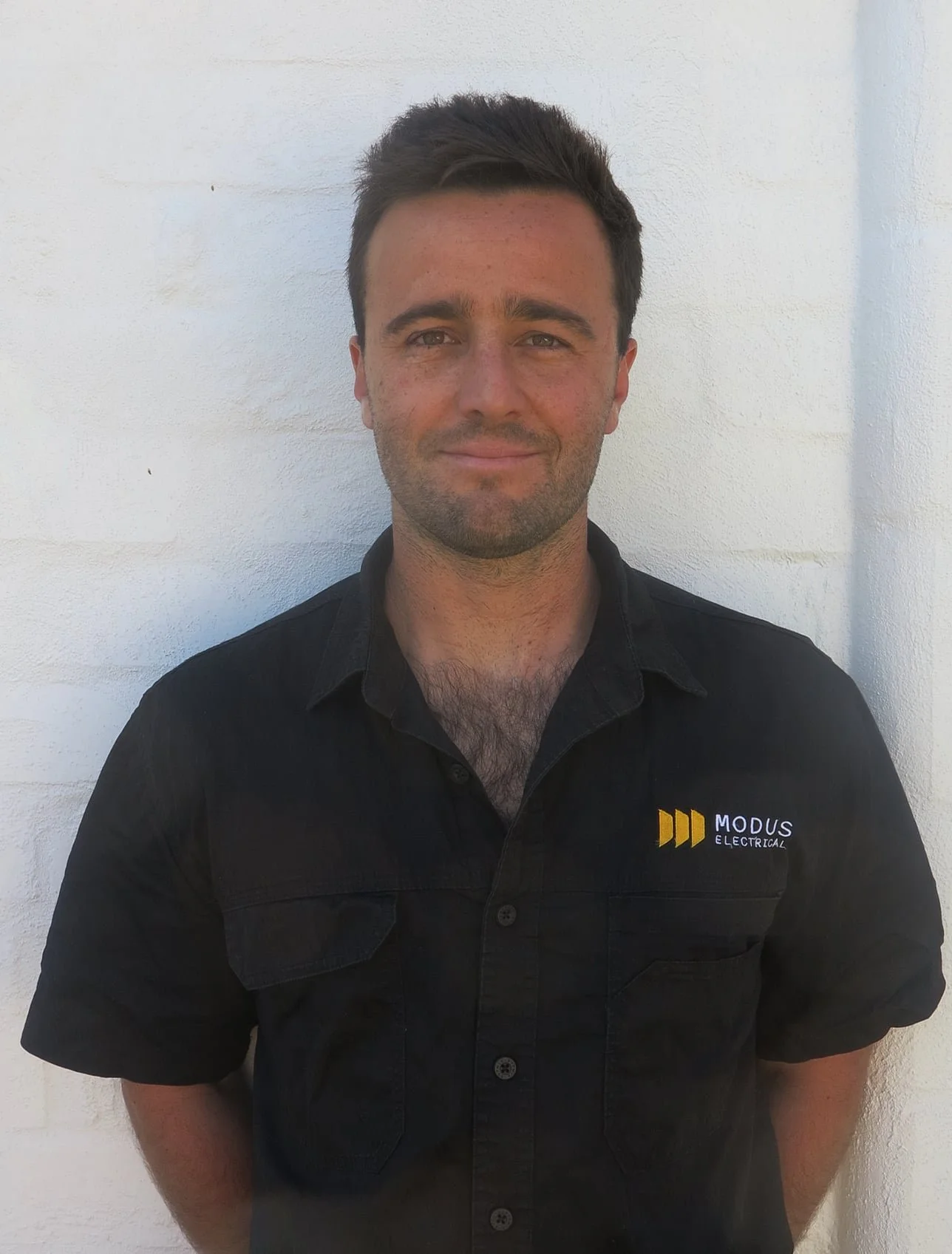 A man with short dark hair and light skin wearing a black shirt with a logo that reads 'MODUS ELECTRICAL,' standing in front of a white brick wall.