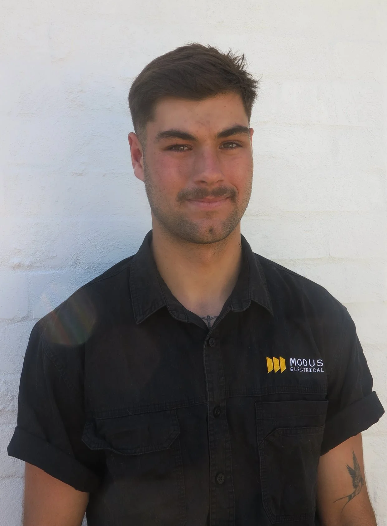 A young man with dark hair, a slight beard, dressed in a black button-up shirt with a logo that says 'MODUS ELECTRICAL,' standing against a white wall, smiling softly at the camera.