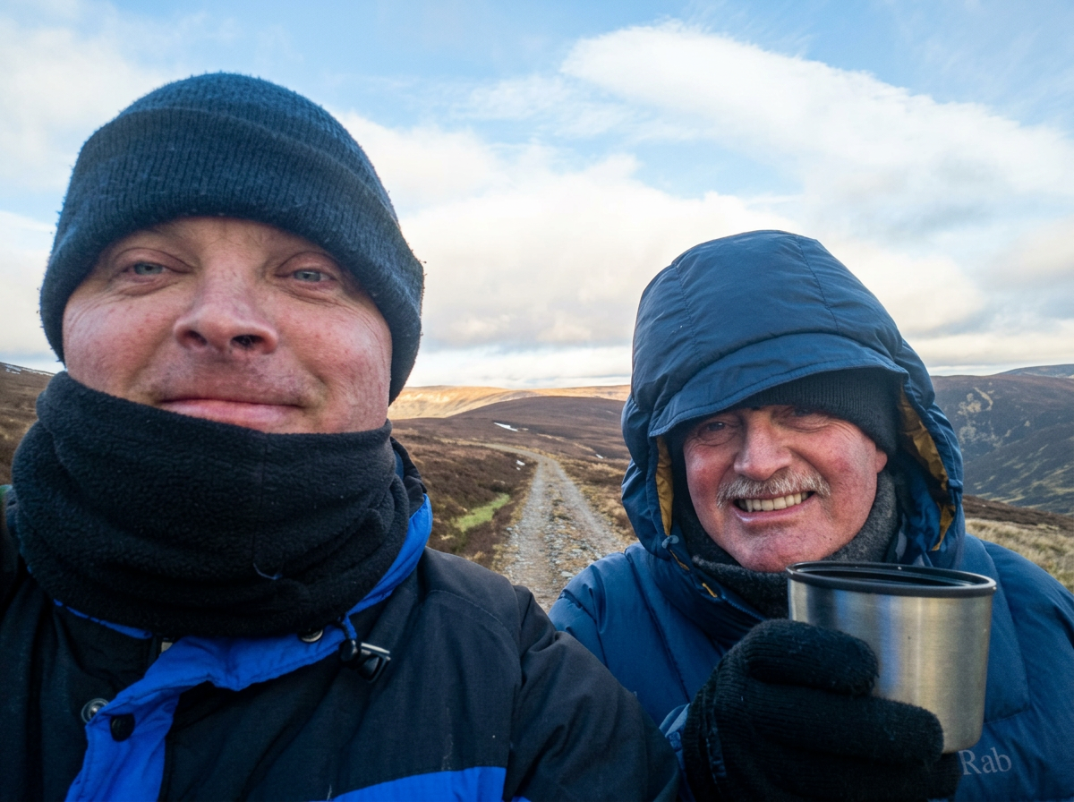 Two men dressed in outdoor winter gear taking a selfie on a mountain trail, with hills and sky in the background.