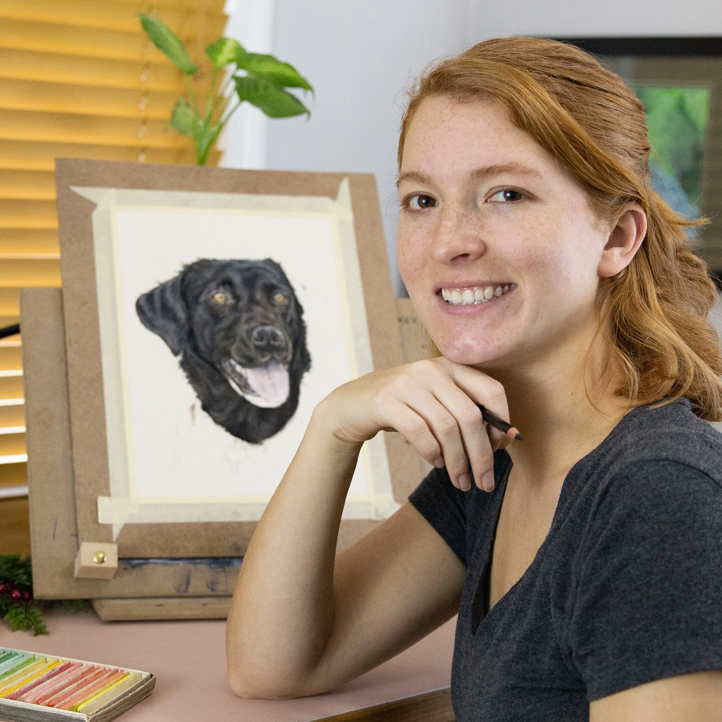 A woman with red hair and freckles smiling and resting her chin on her hand sits at a table with colored chalk and an art piece of a black dog in the background.